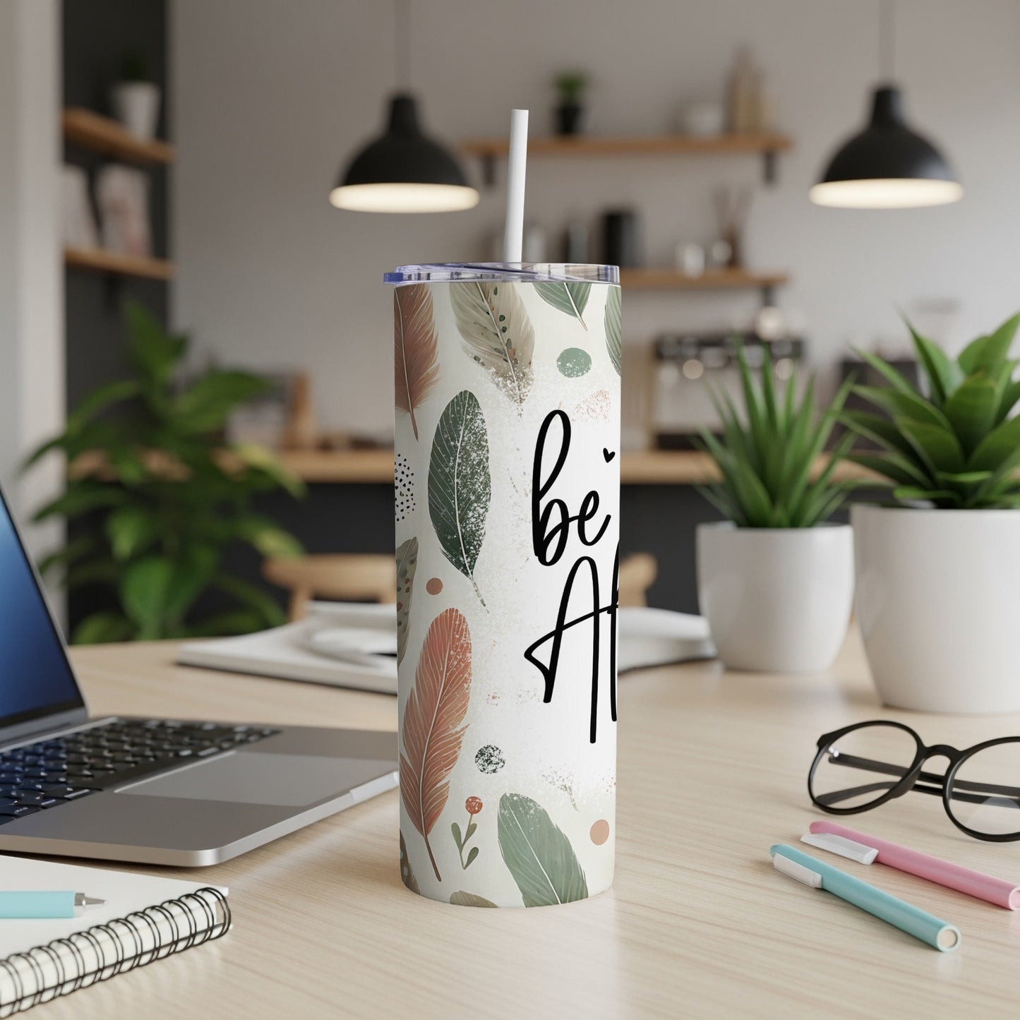 Tall tumbler with feather pattern and "be kind" text, on a desk.