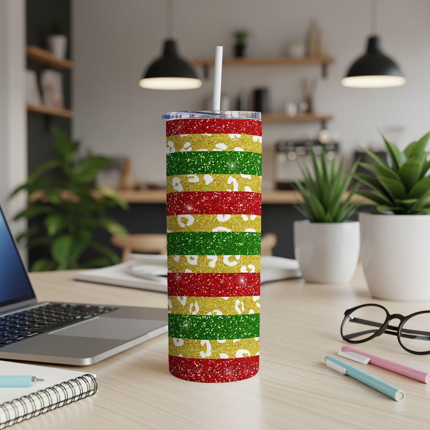 Glittery red, green, and gold striped tumbler with straw on a desk.