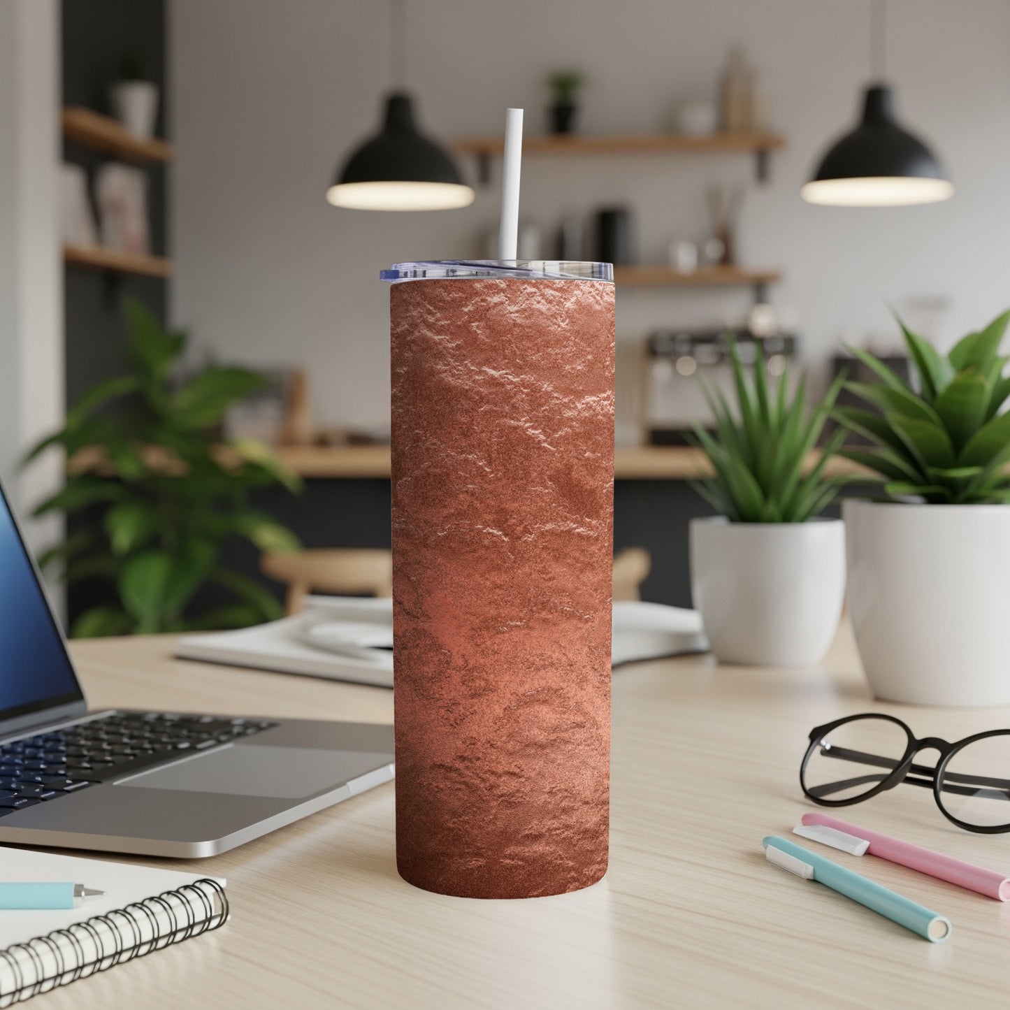 Copper textured tumbler with a straw on a wooden desk with a laptop and glasses.