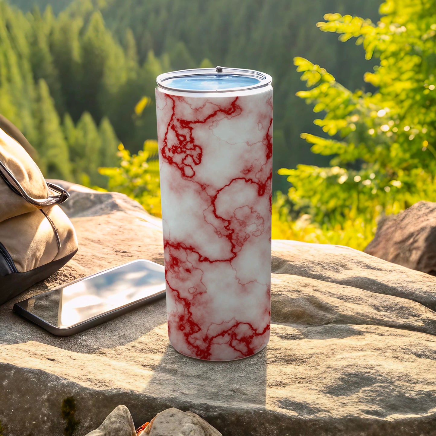 Red marble pattern tumbler on rock with phone and bag.