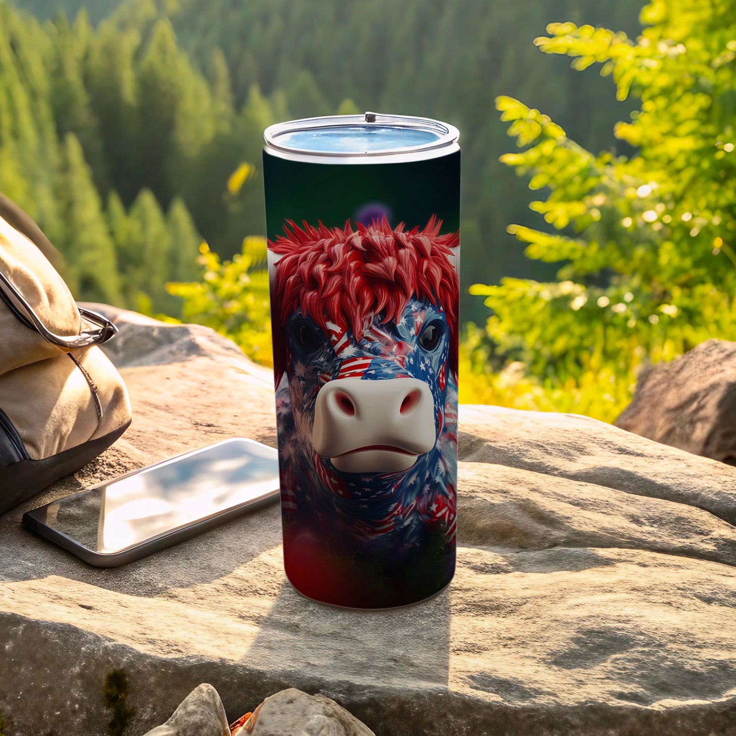 Tumbler with patriotic cow design sits on rocks with phone and bag.