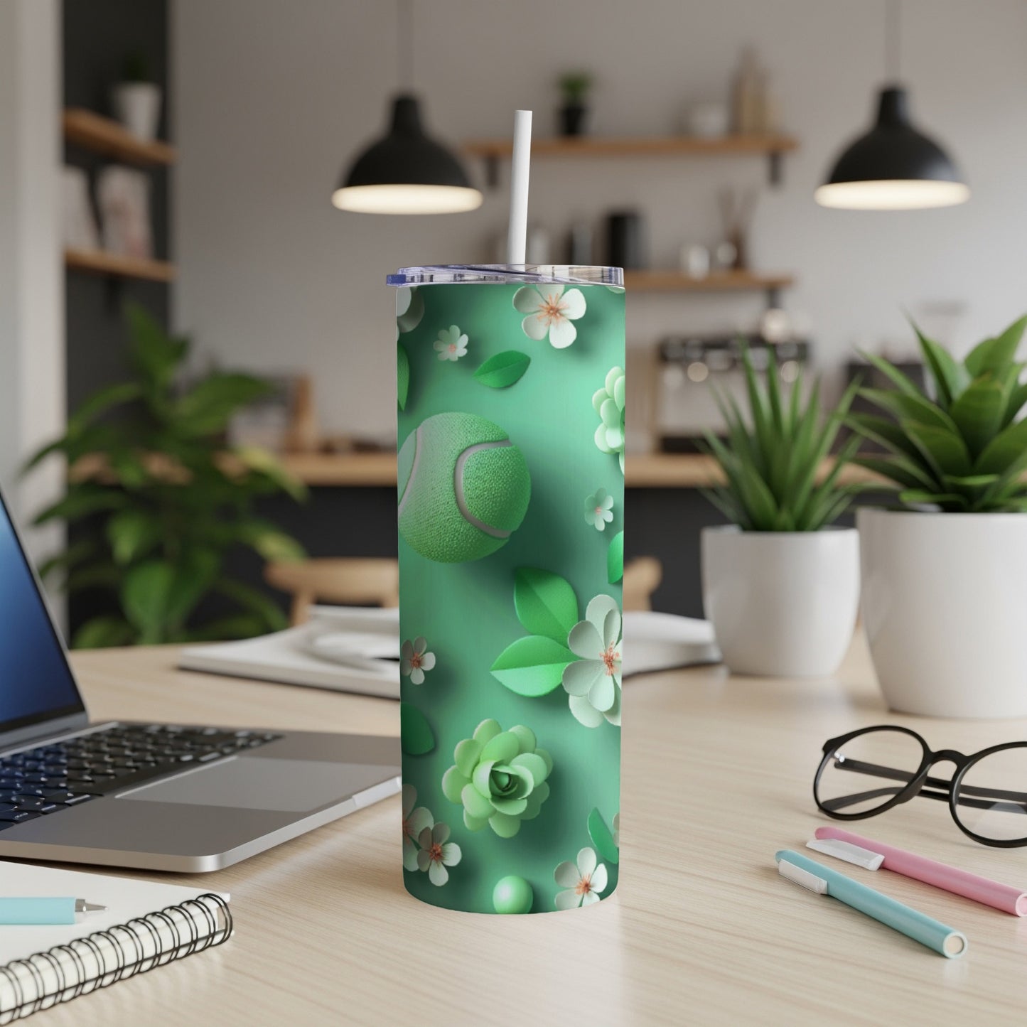 Green tumbler with floral and tennis ball pattern, straw, laptop, and pens.