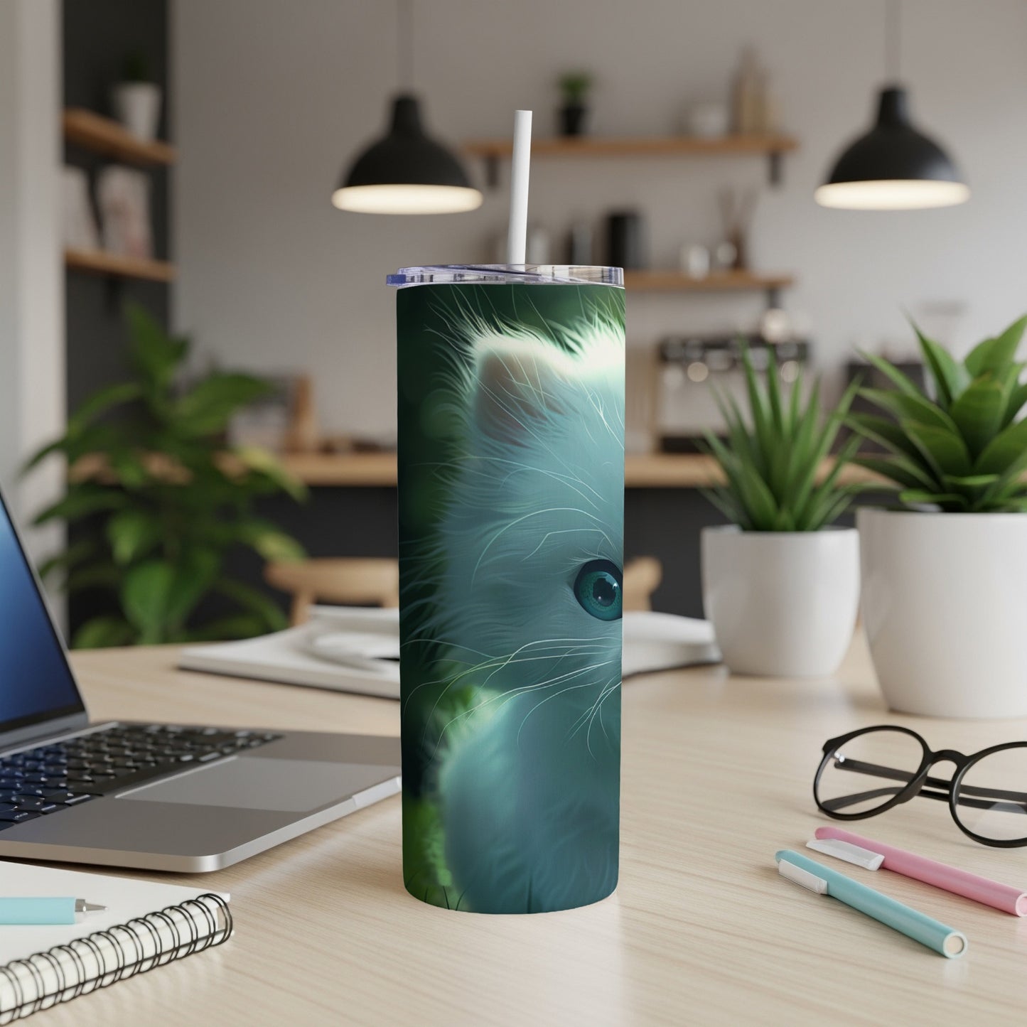 White kitten face on a skinny tumbler with straw, laptop and pens nearby.