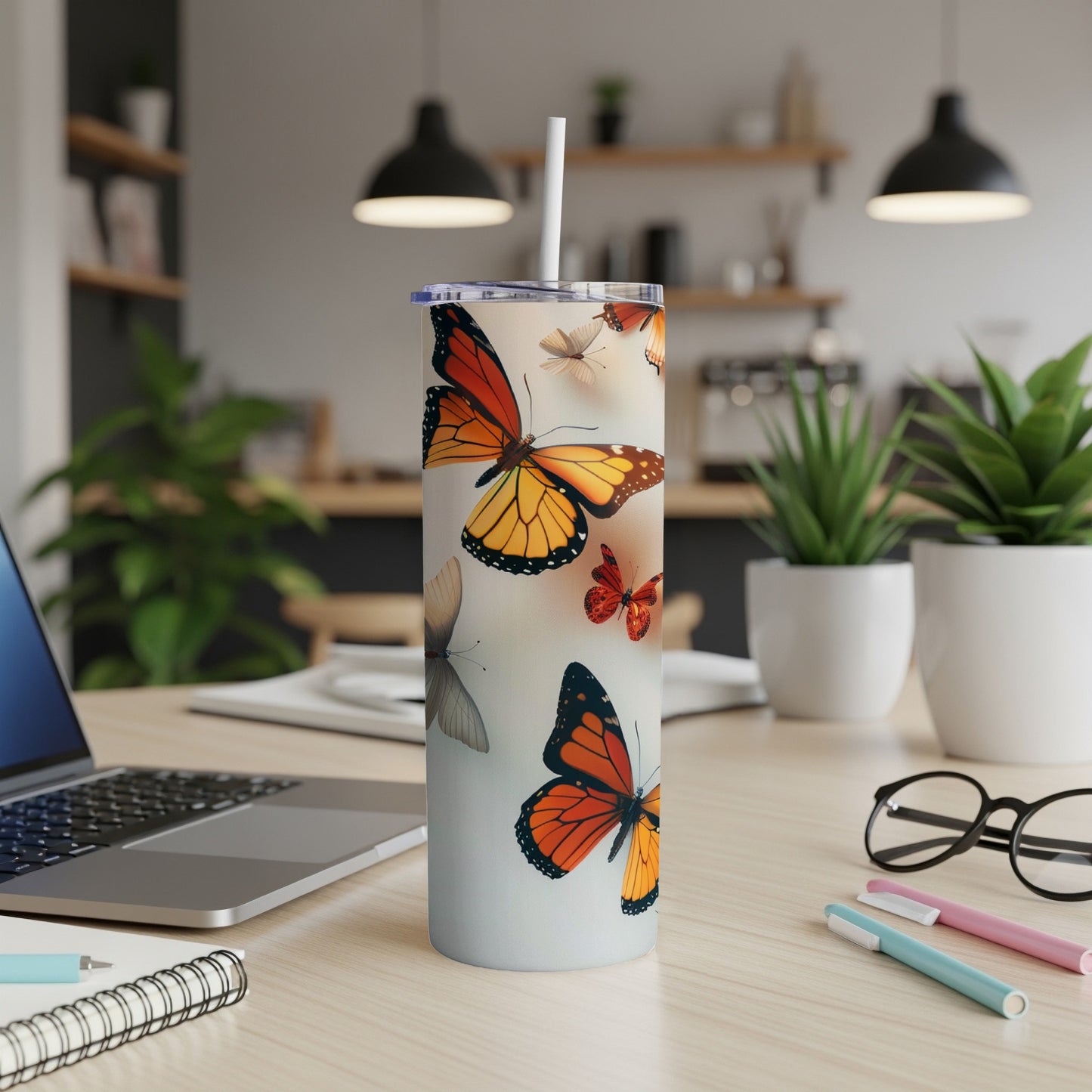 Tall tumbler with monarch butterflies, straw, laptop, and pens on a desk.