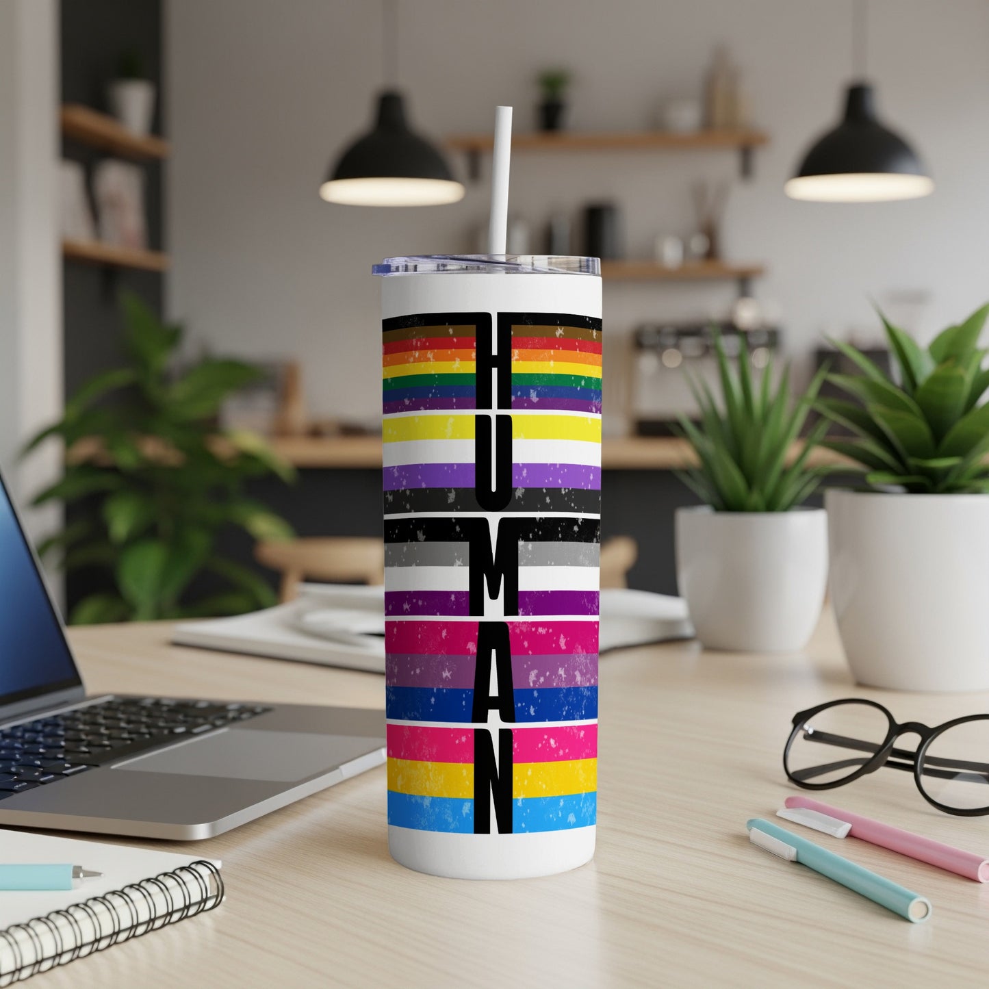 Pride flag tumbler with "HUMAN" text, laptop, and plants in background.