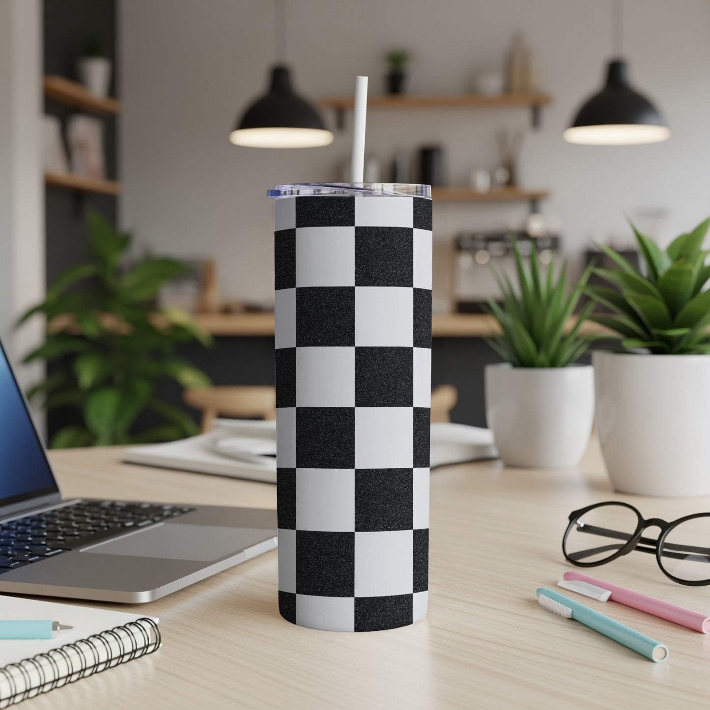 Checkerboard pattern tumbler with a straw on a desk next to a laptop.