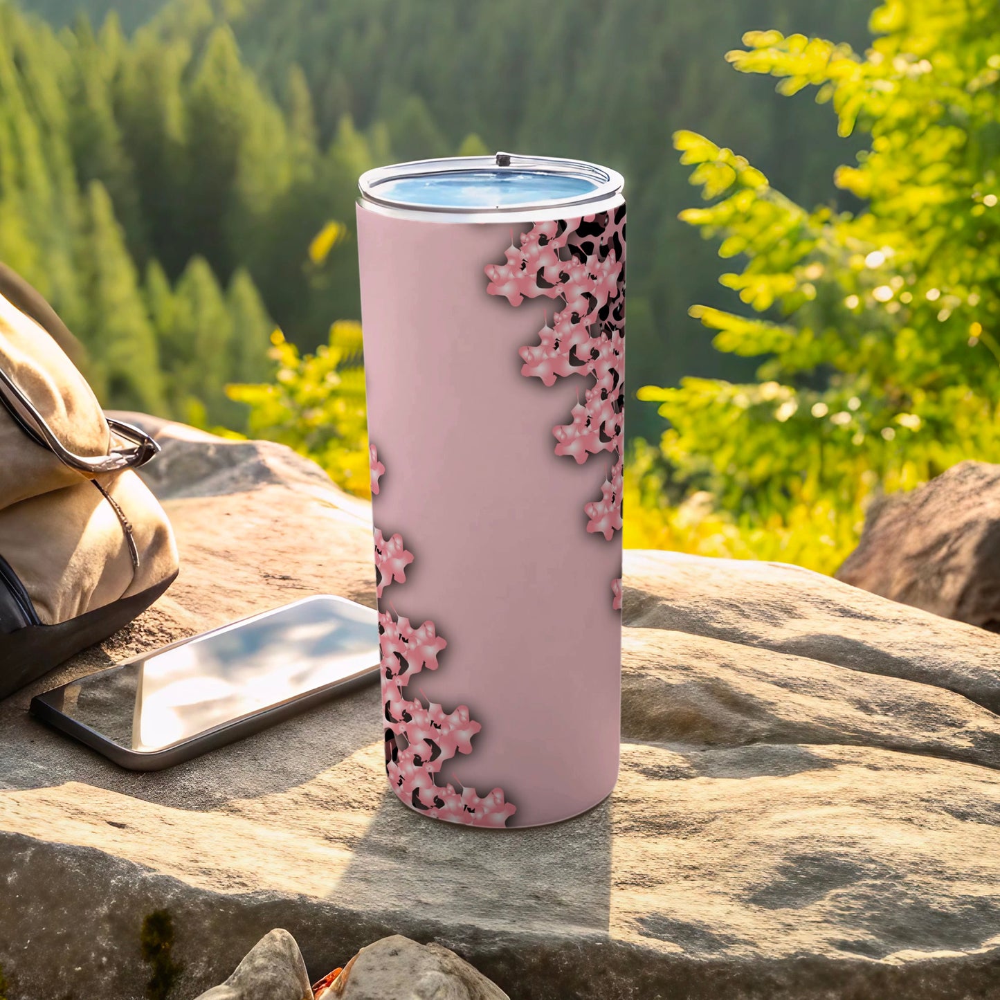 Pink tumbler with floral pattern on rocks with phone and backpack.