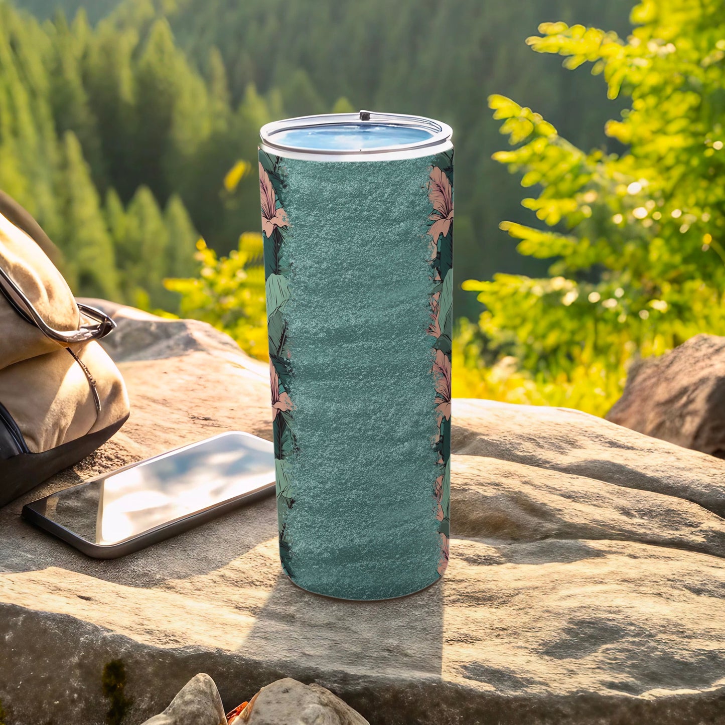 Teal tumbler with floral border, on rocks, with backpack and phone.