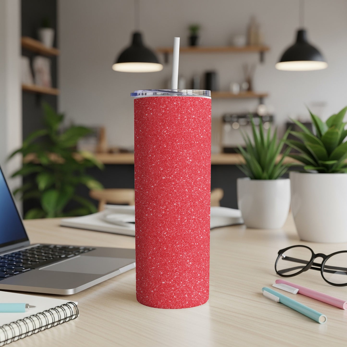 Red glitter tumbler with straw on a wooden desk near a laptop and plants.