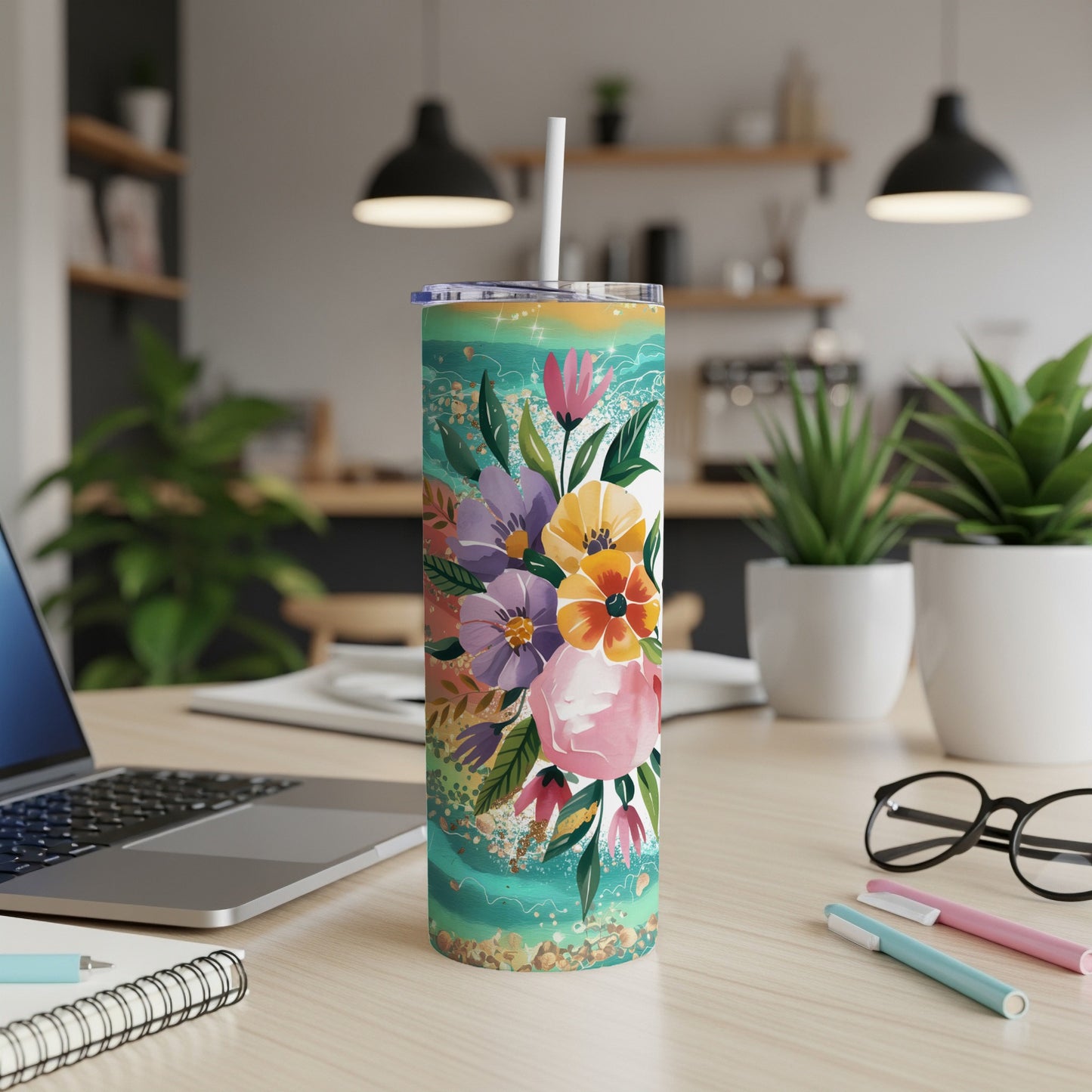 Floral tumbler with straw on desk beside laptop and pens.