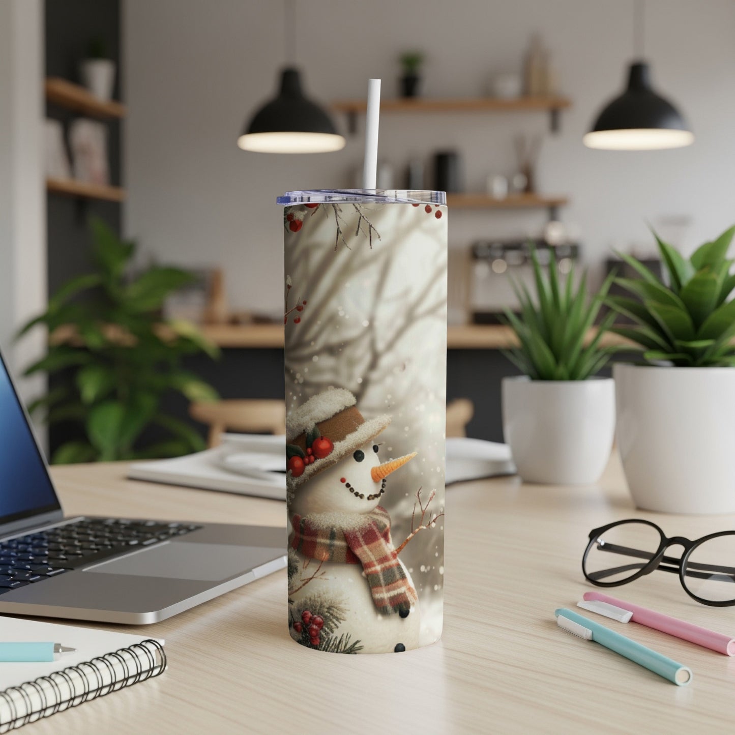 Snowman tumbler with plaid scarf and hat on desk with laptop and plants.