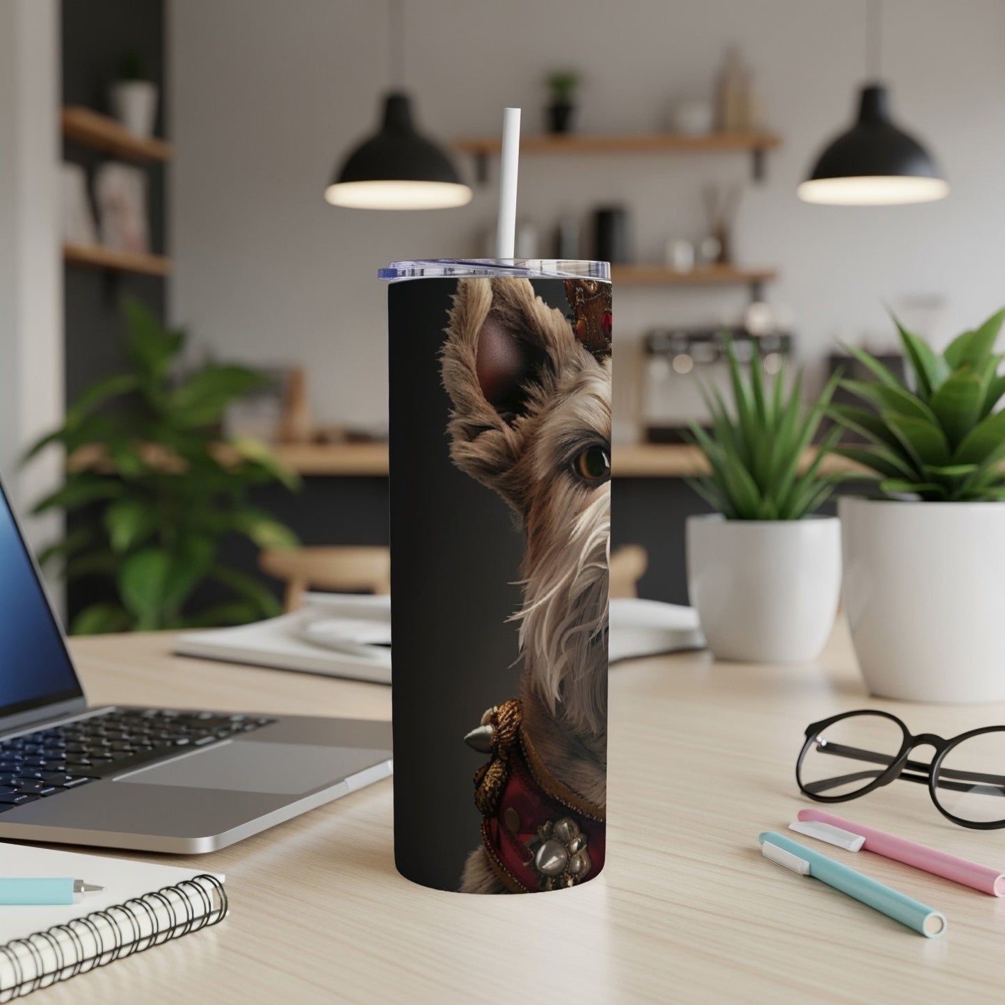Dog with crown on a tumbler, on a desk with laptop and pens.