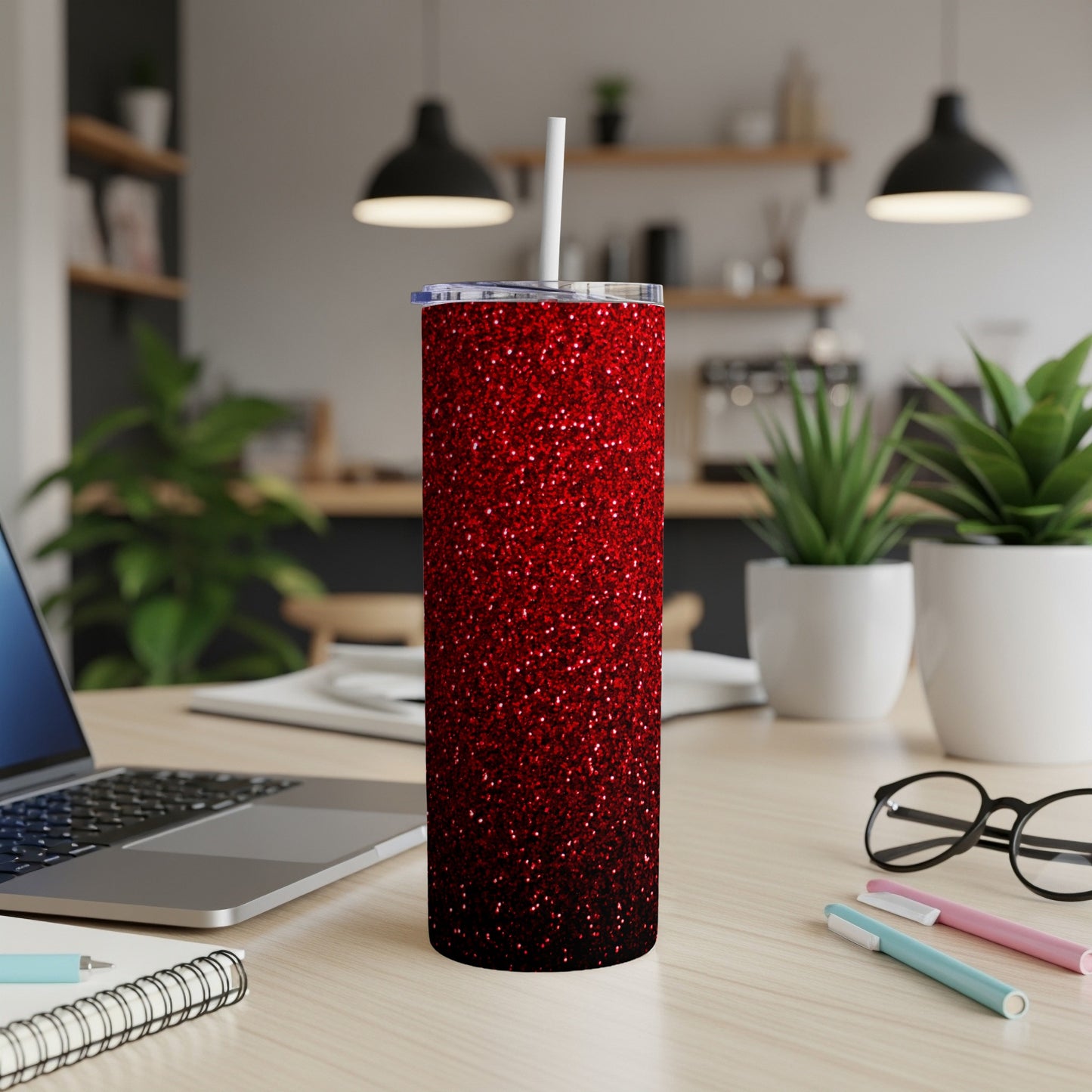 Red glitter tumbler with straw on desk, laptop and plants in background.