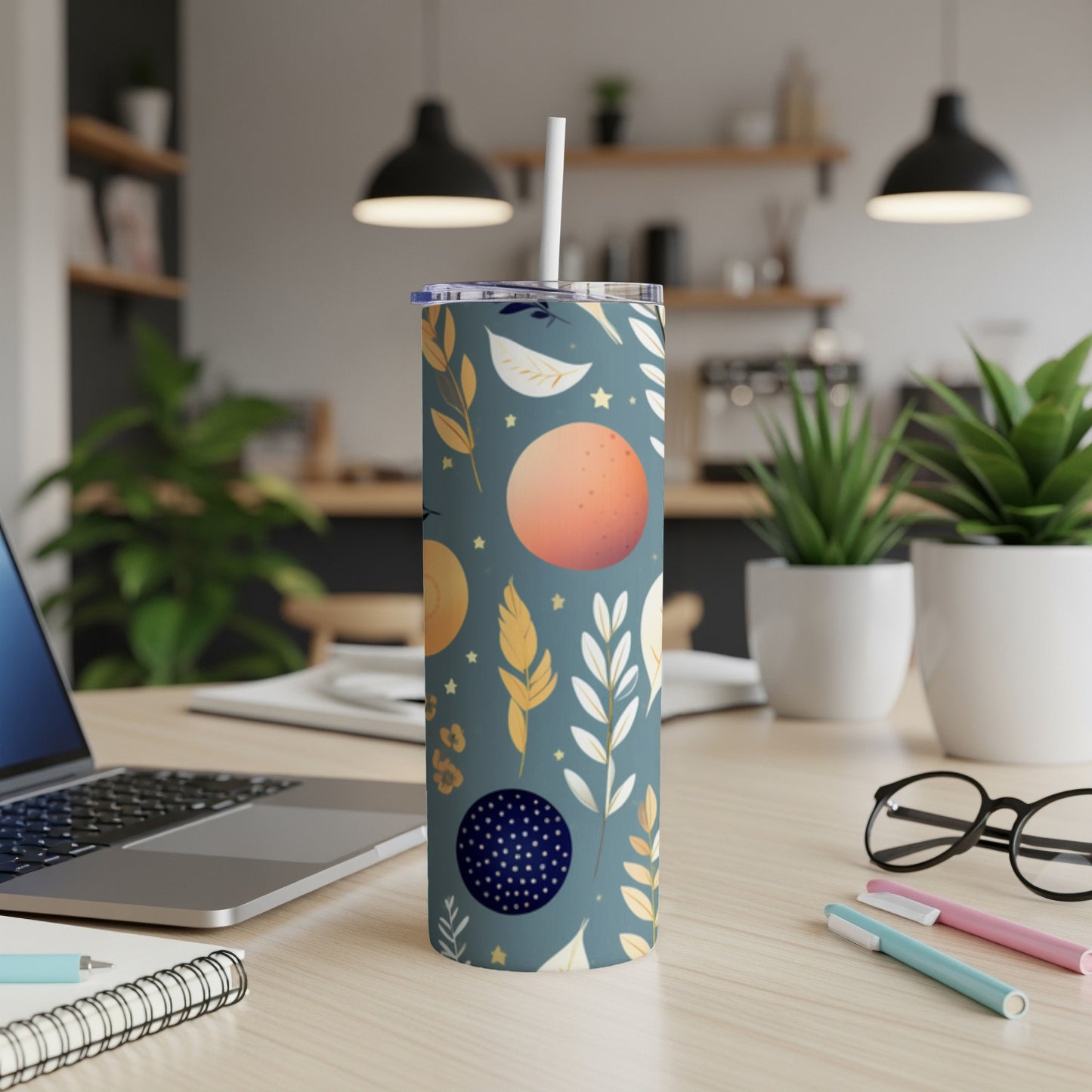 Tall teal tumbler with moon and leaf pattern, straw, on desk.