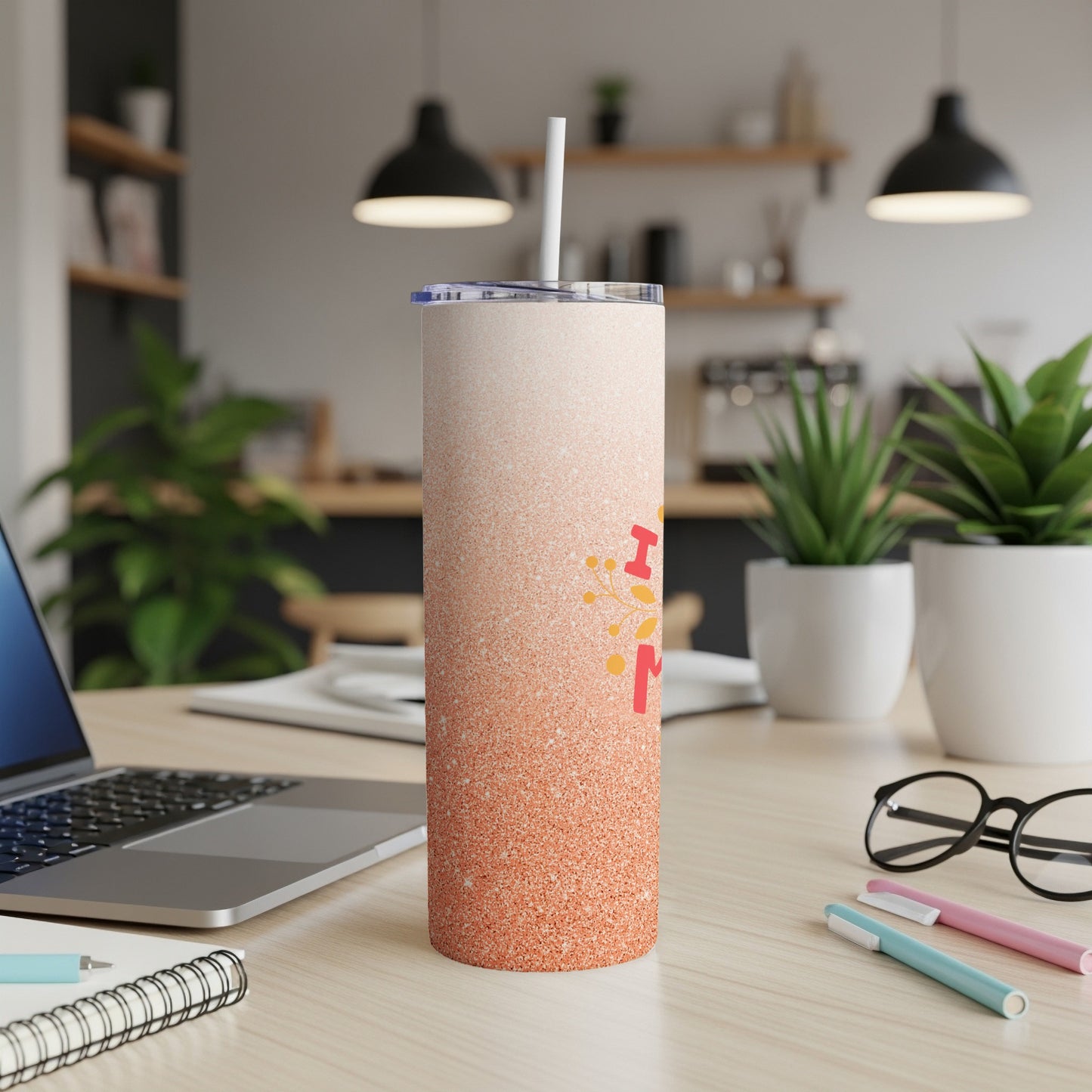 Glittery orange tumbler with "Mom" text on a desk with a laptop and pens.