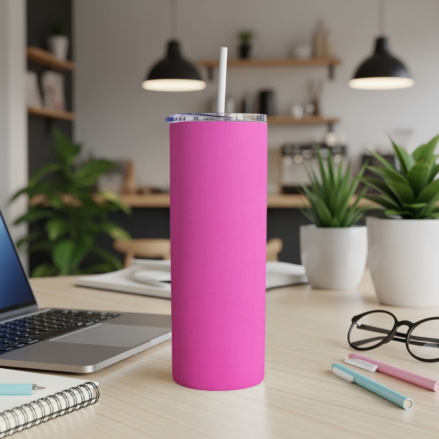 Pink tumbler with white straw on a desk with laptop and pens.
