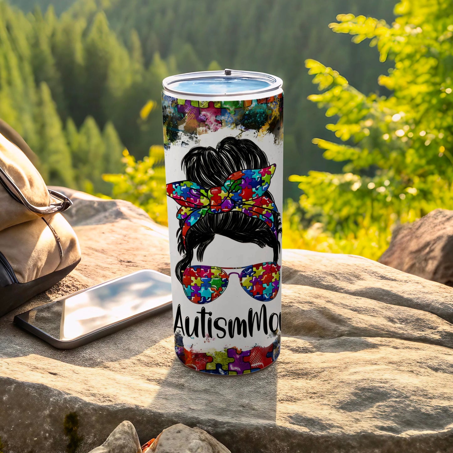 Tumbler with Autism mom design, puzzle pieces, messy bun, sunglasses, on a rock.