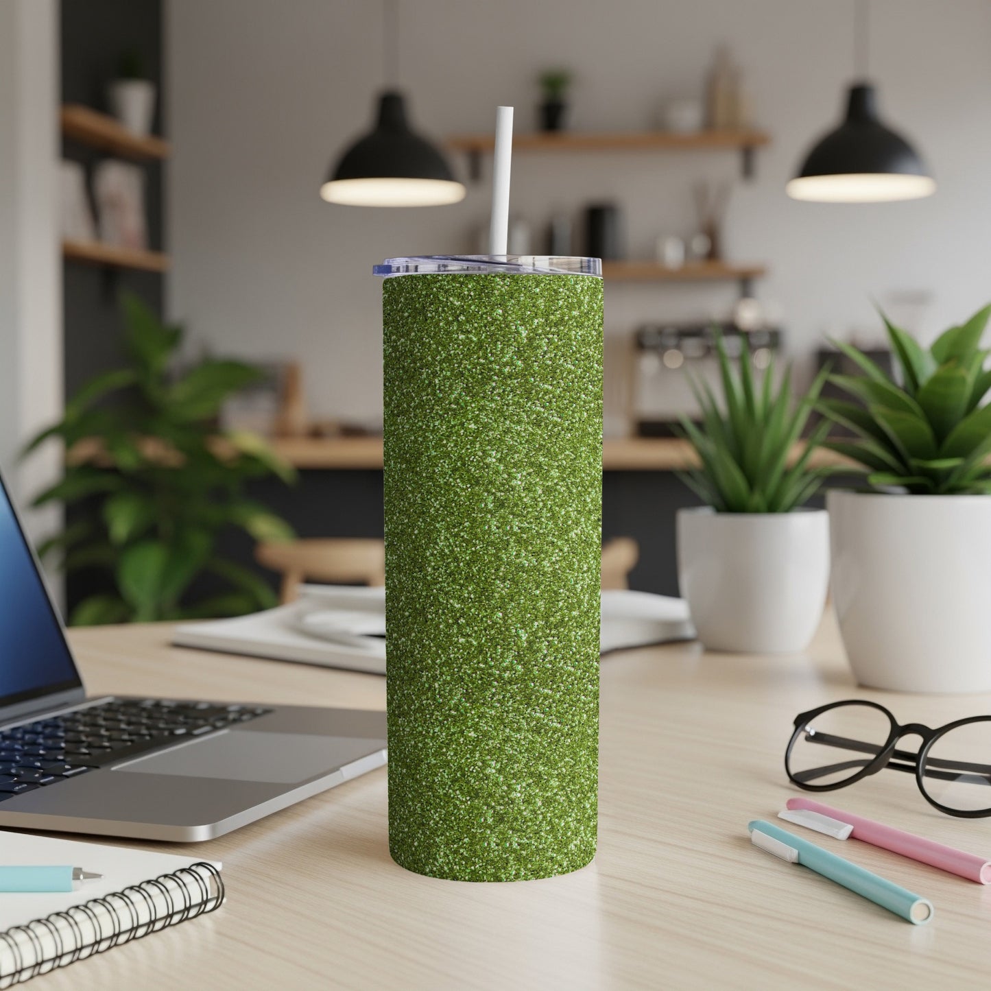 Green glitter tumbler with straw on a wooden desk with laptop and supplies.