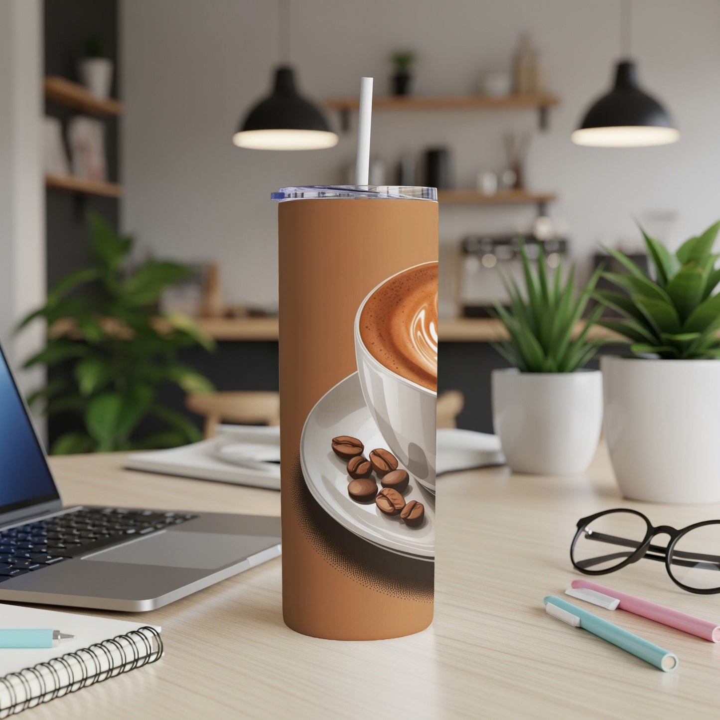 Brown tumbler with latte art and coffee beans, laptop, and pens.