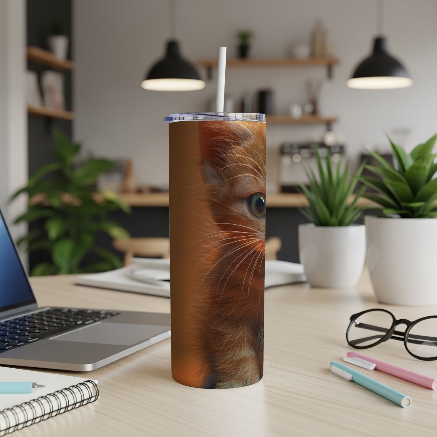 Orange tabby kitten peeking from behind a white tumbler on a desk.