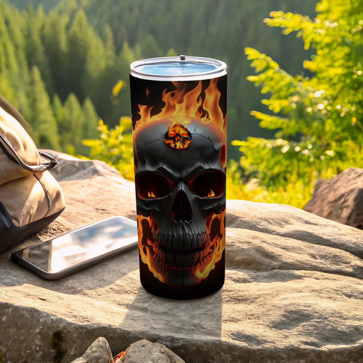 Black tumbler with flaming skull design sits on rocks with phone and backpack.