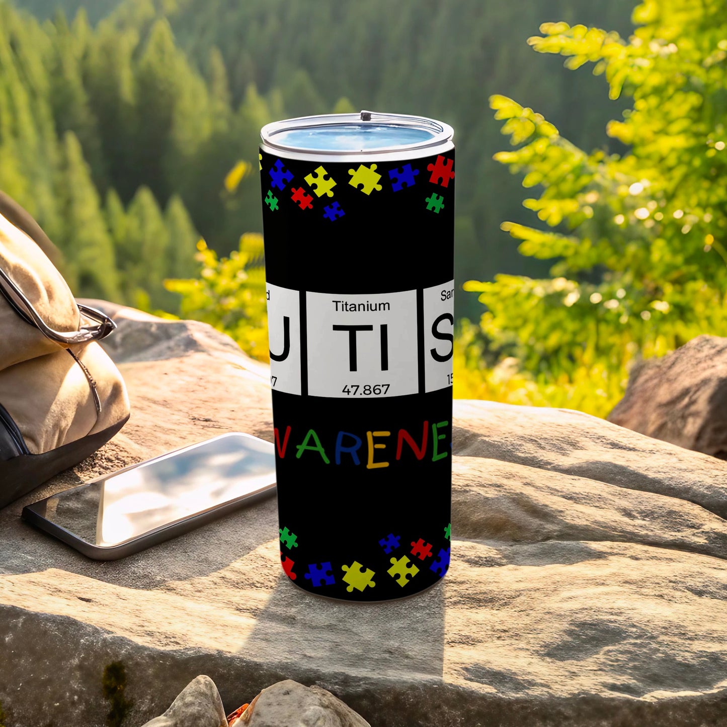 Tumbler with "AUTISM AWARENESS" text and puzzle pieces, on rocks.