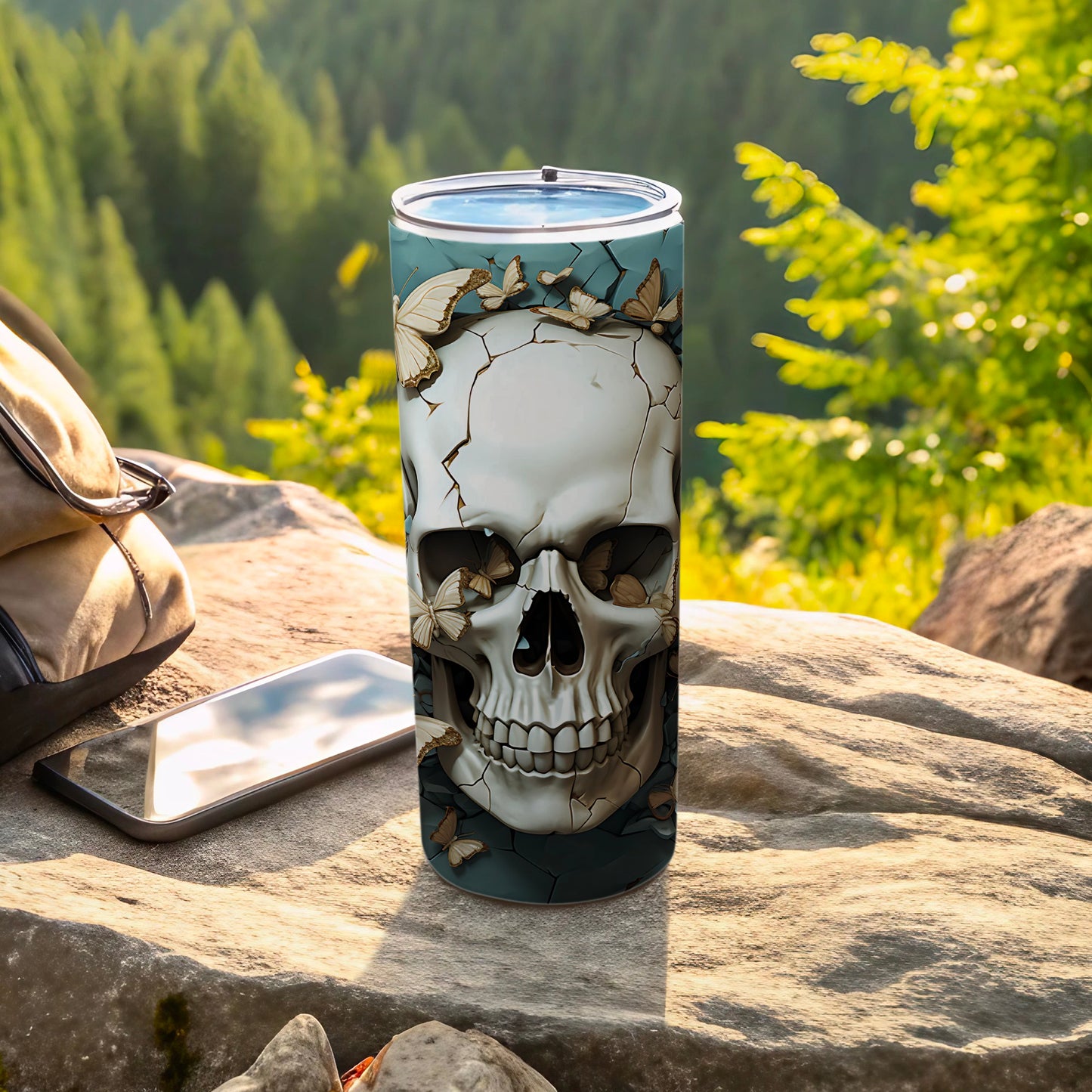 Skull tumbler with butterflies, beside a phone and backpack on rocks.