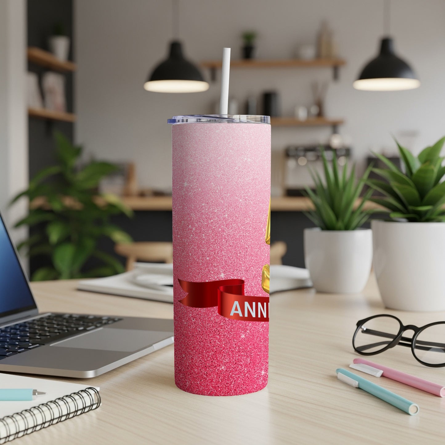 Pink glitter tumbler with "Anniversary" text on a desk with a laptop and plants.
