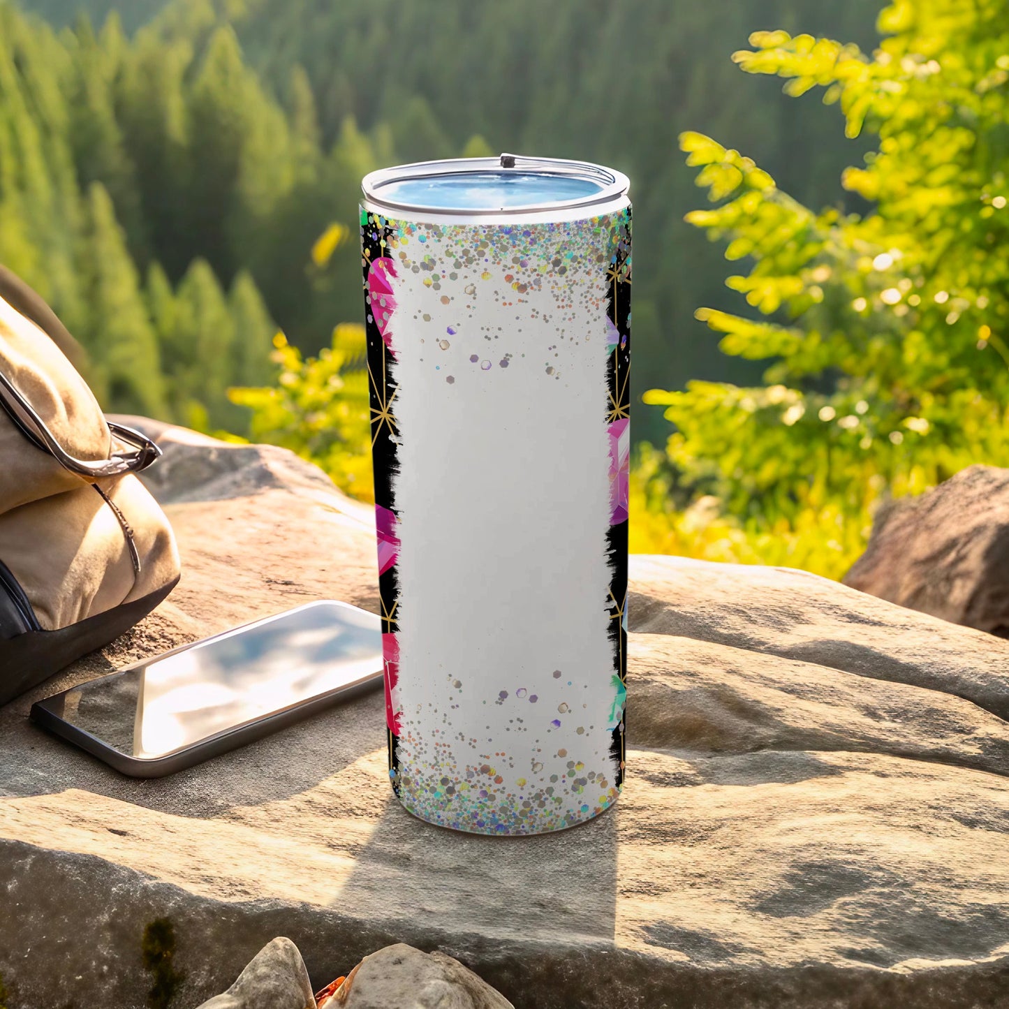 Glittery floral tumbler on rock with backpack and phone outdoors.