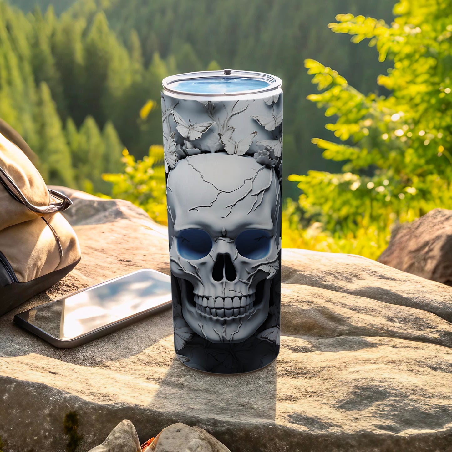 Skull tumbler with blue eyes and floral accents on rocky surface with phone and backpack.