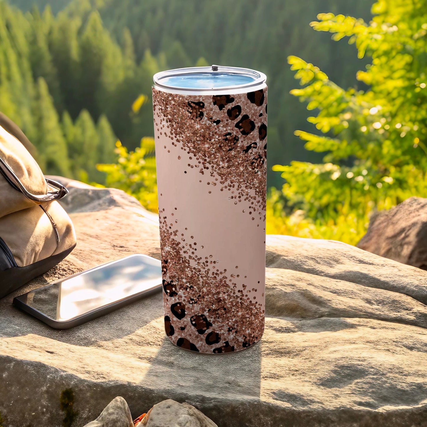 Pink tumbler with leopard print and glitter on a rock with phone and backpack.