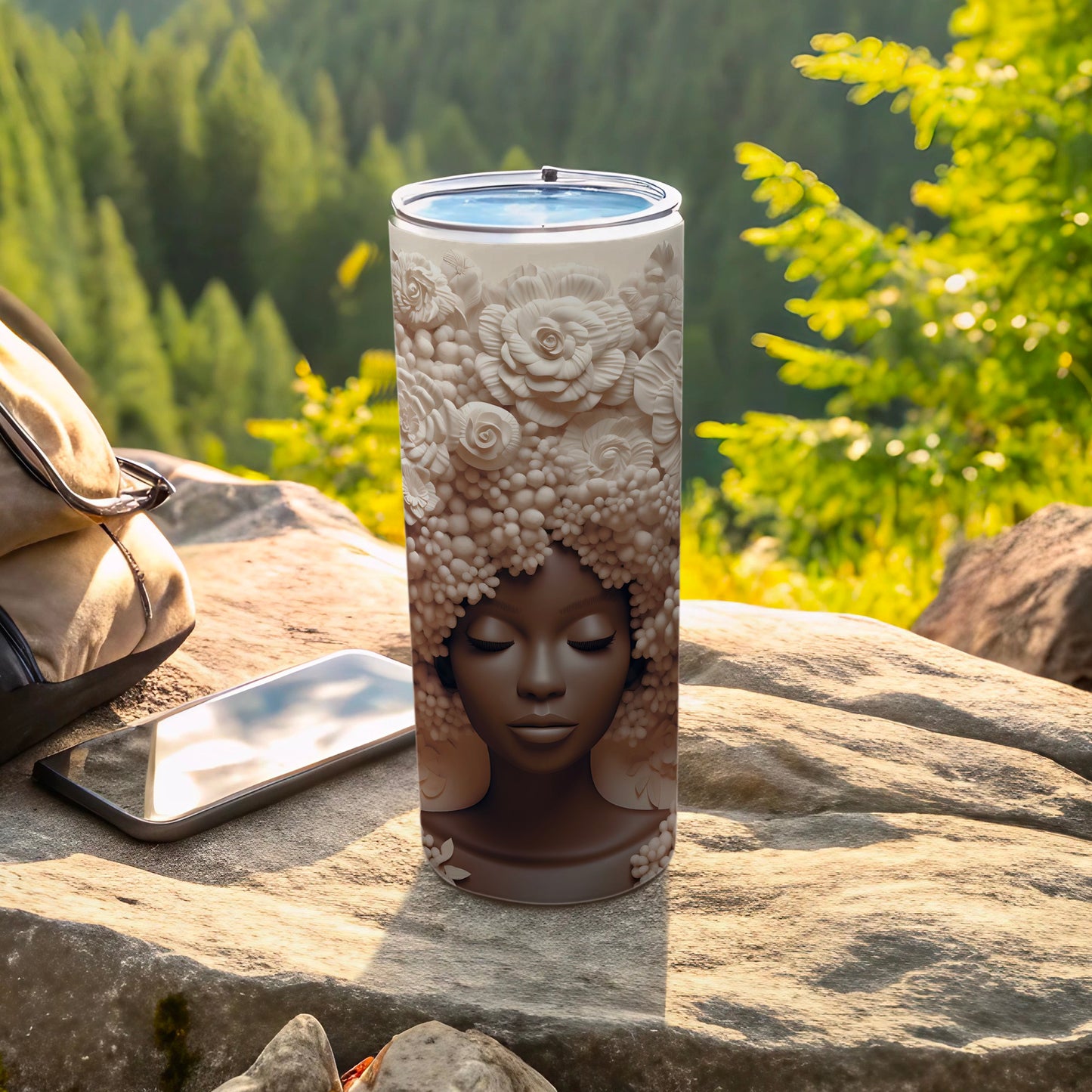 Tumbler with woman's face and floral headdress, rests on rocks with phone and bag.