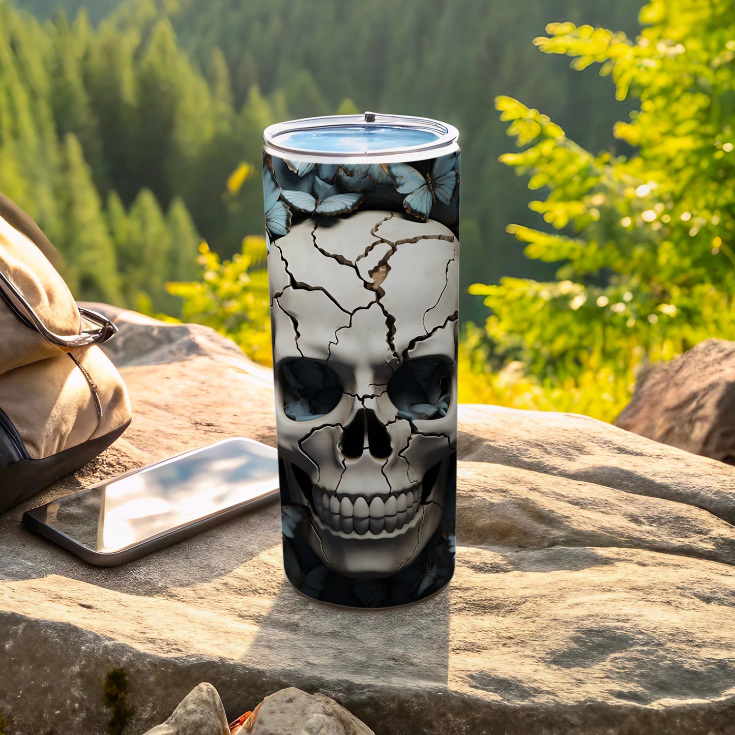 Skull tumbler with butterflies and cracks, resting on rocks outdoors.