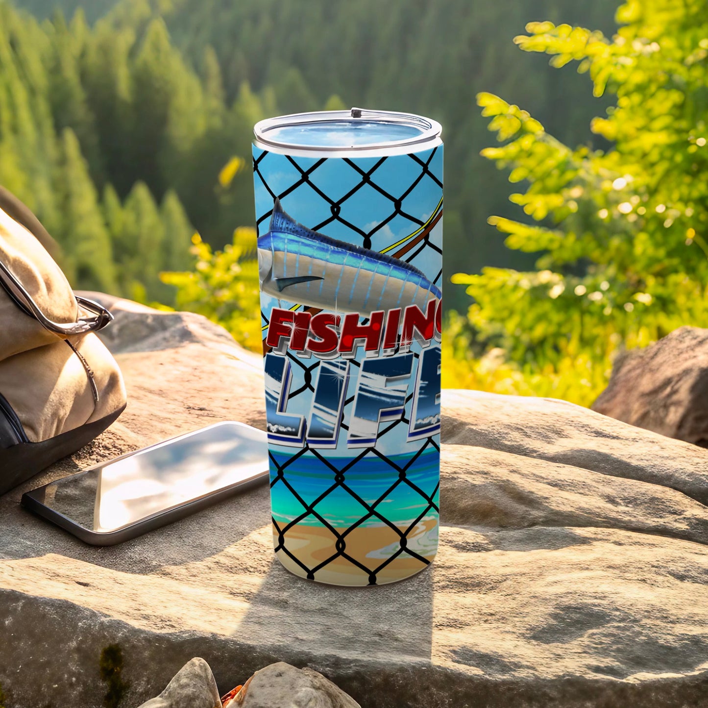 Fishing Life tumbler with a marlin graphic on a rock by a backpack and phone.