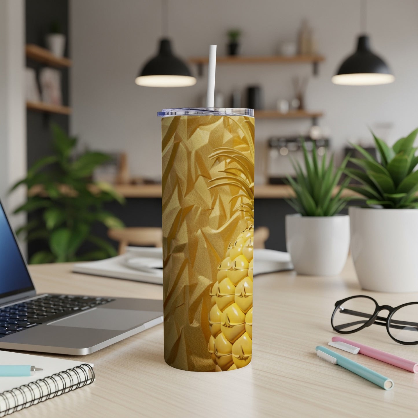 Golden pineapple tumbler with straw on a desk next to a laptop and pens.
