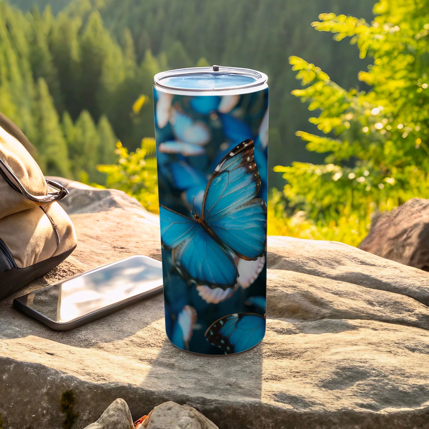 Blue butterfly print stainless steel tumbler on rocky surface with phone and backpack.