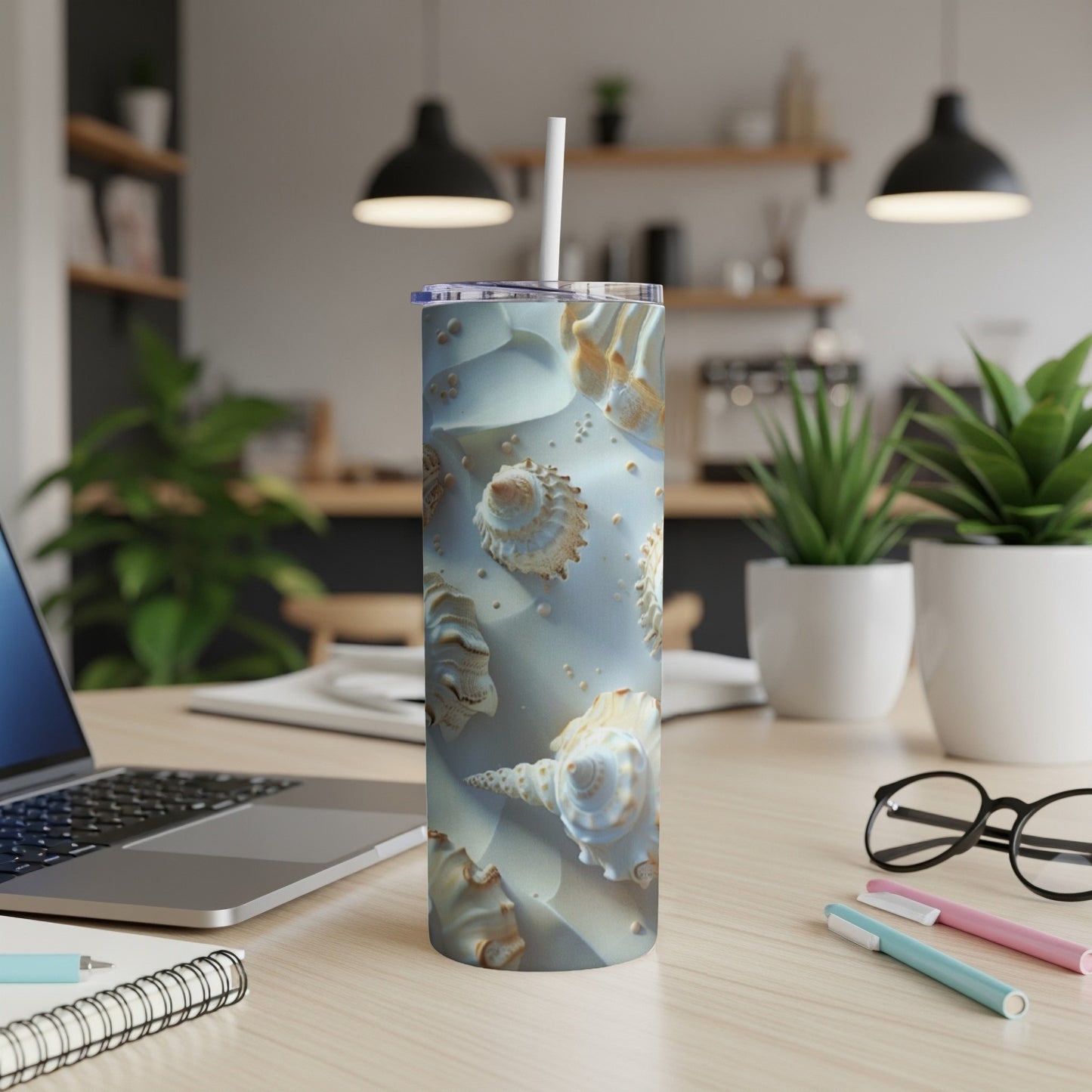 Tall tumbler with seashell design on a desk with laptop and pens.