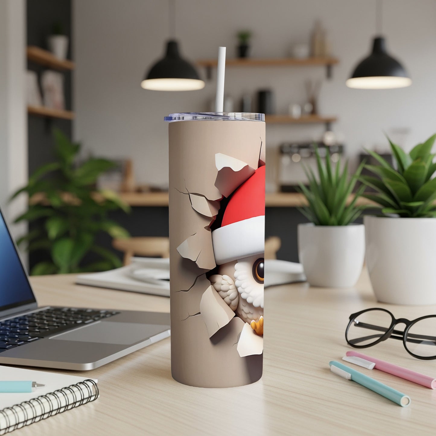 Owl in Santa hat peeking through cracked tumbler, on desk with laptop and pens.
