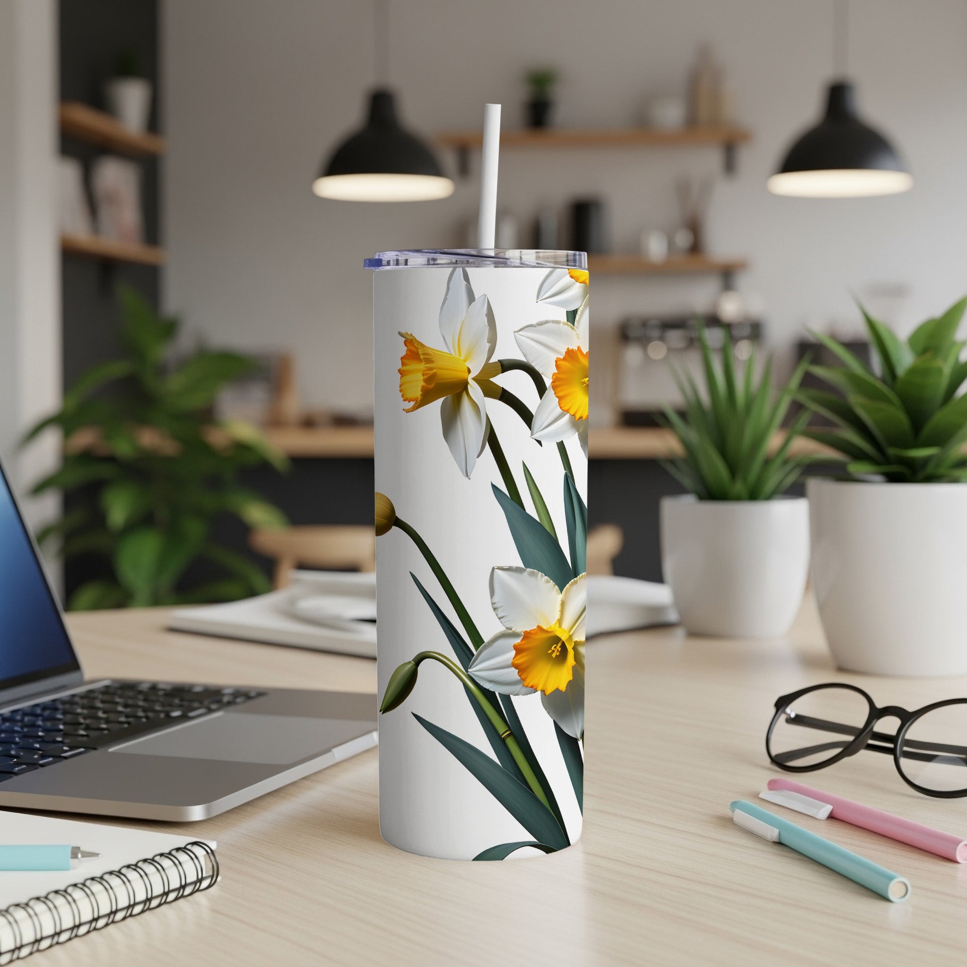 White tumbler with daffodils and green leaves, on a desk.