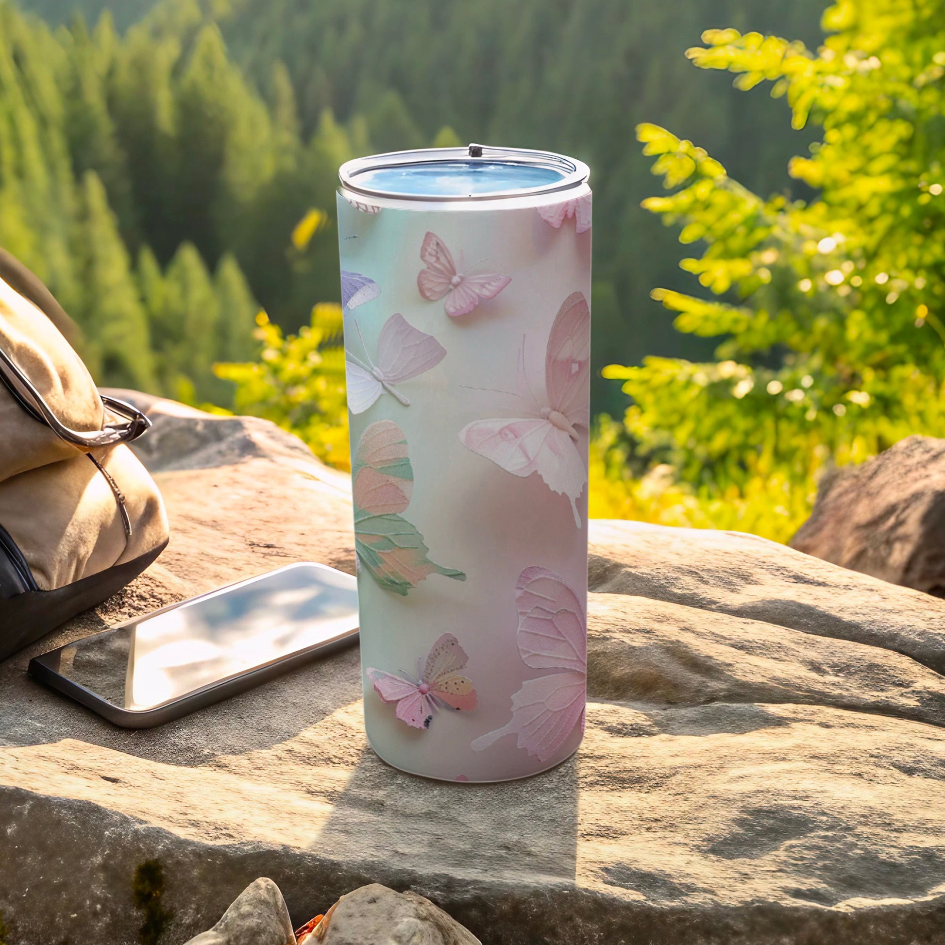 White tumbler with pastel butterflies, next to phone and bag on rock.