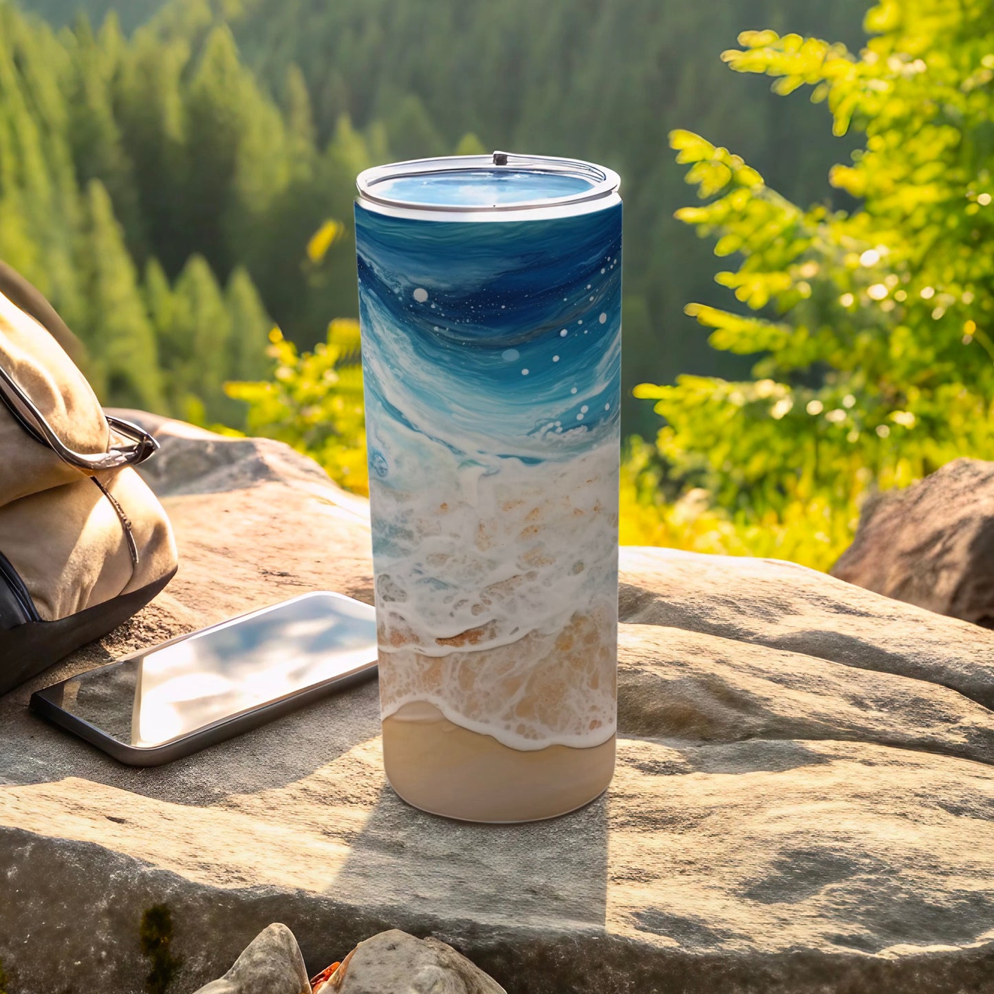Tall tumbler with ocean wave design on a rock, with phone and bag.