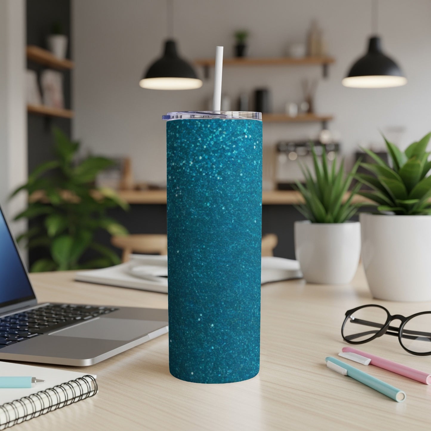 Teal glitter tumbler with straw on a desk with laptop and plants.
