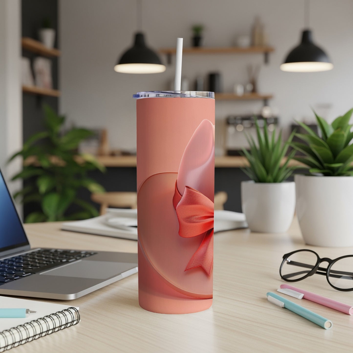 Pink tumbler with red ribbon detail, shown on a desk with a laptop and pens.