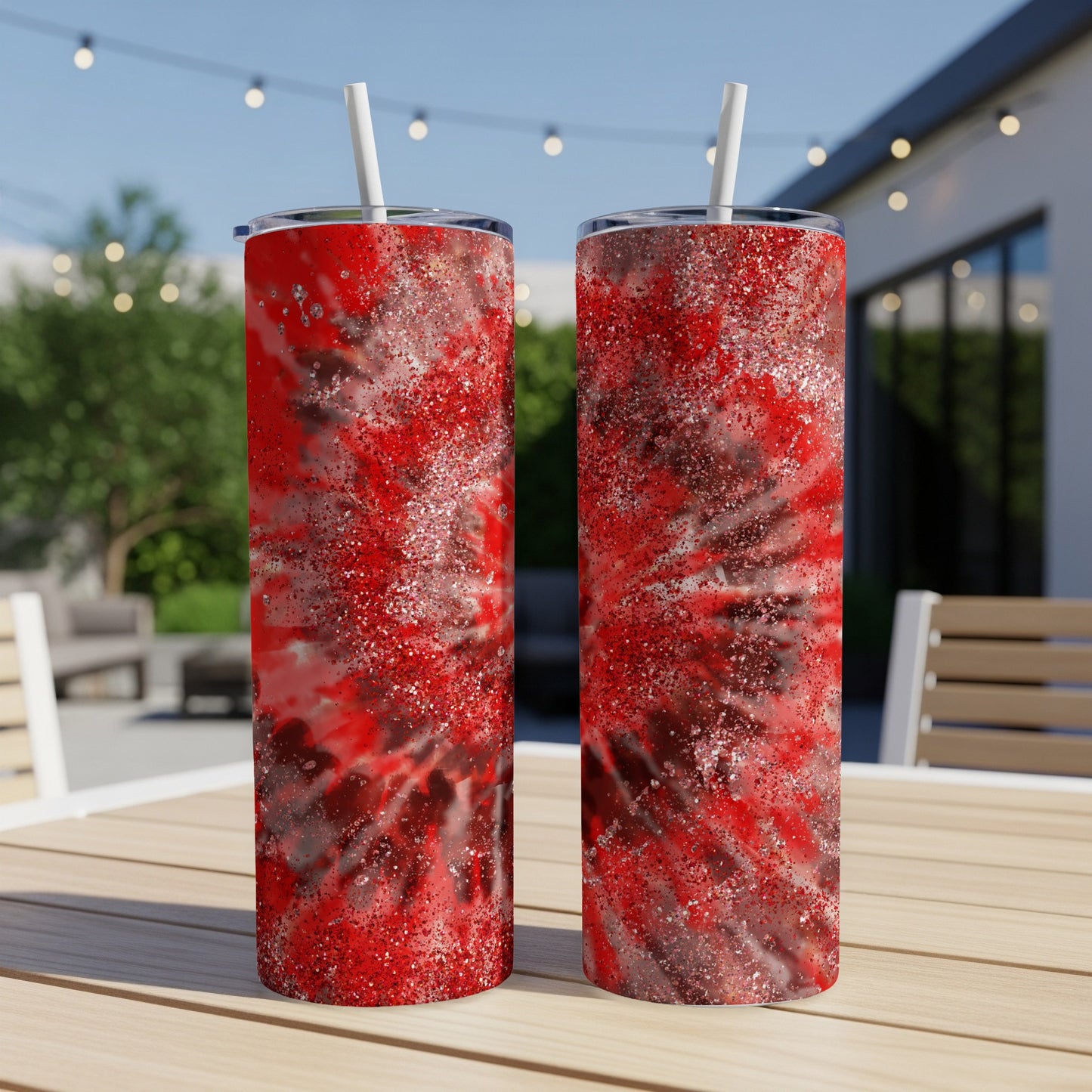 Two red tie-dye tumblers with glitter sit on a wooden table outdoors.