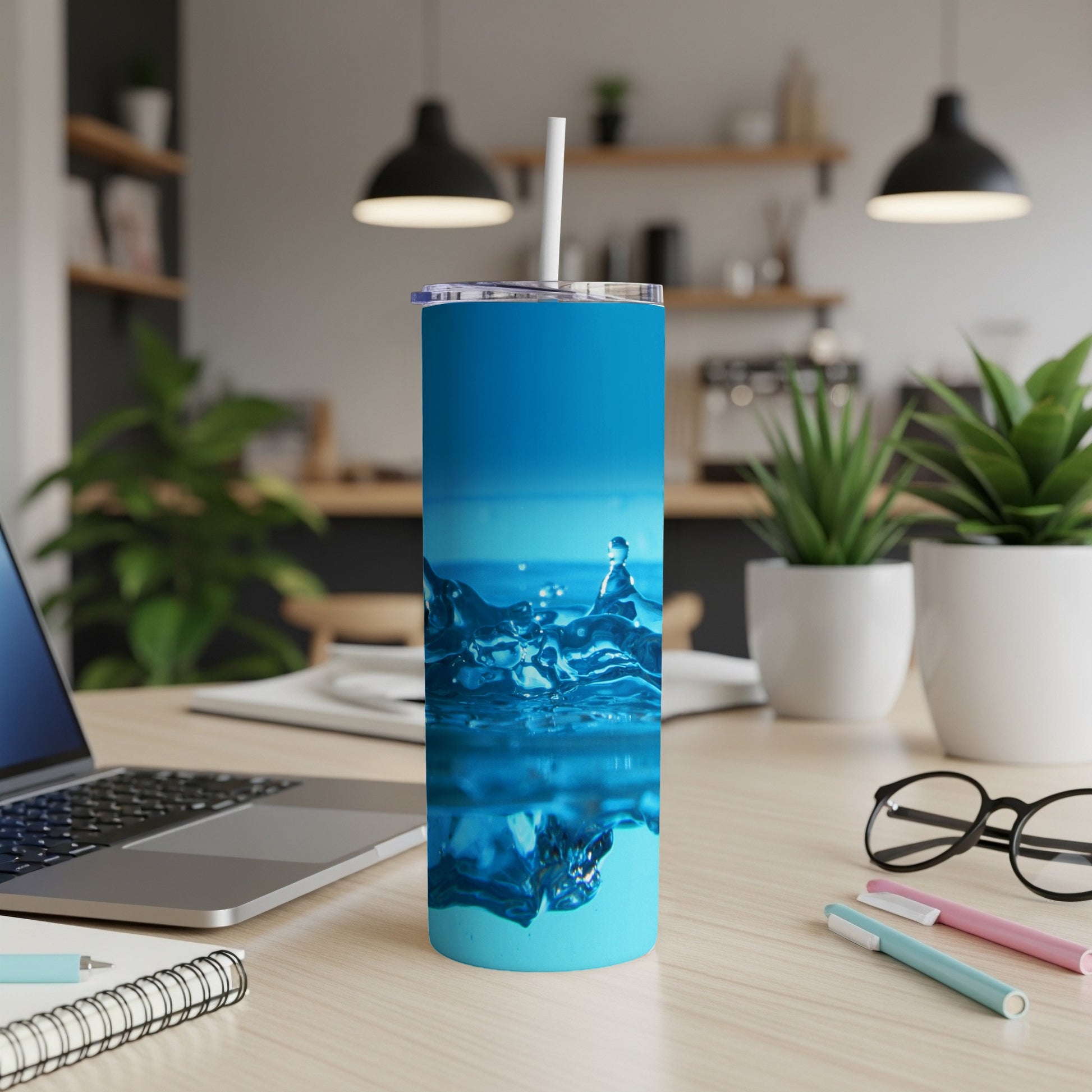Blue insulated tumbler with white straw on desk next to laptop and pens.