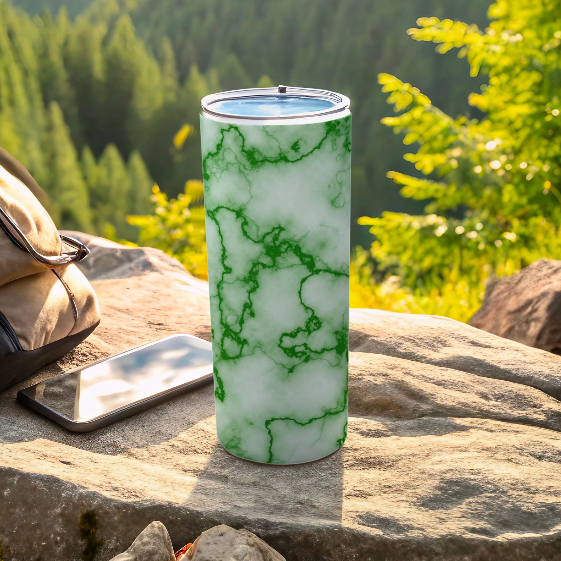 White tumbler with green marble pattern, smartphone, and backpack on rock.
