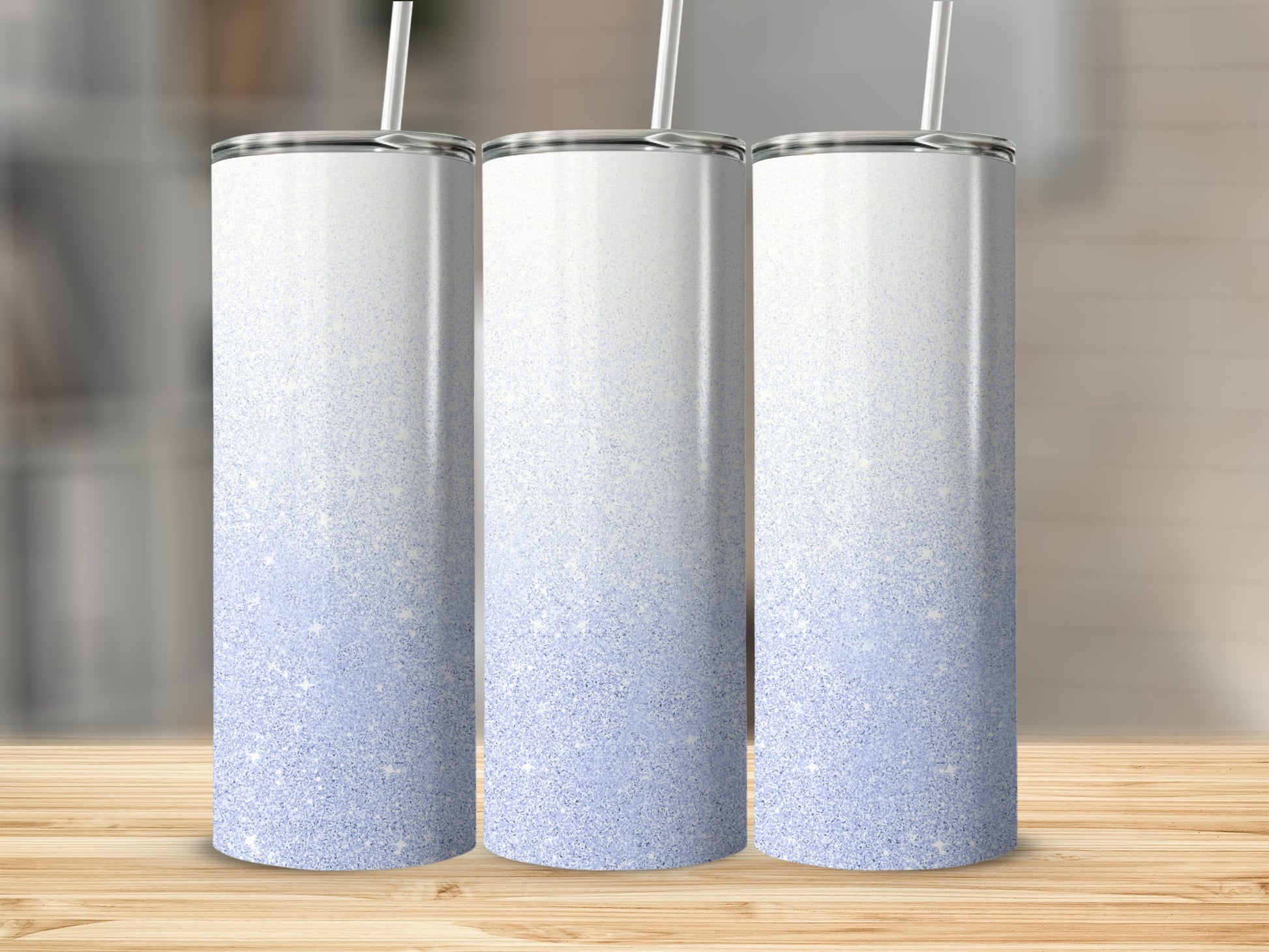 Three glittery white to light blue gradient tumblers with straws on a wooden surface.
