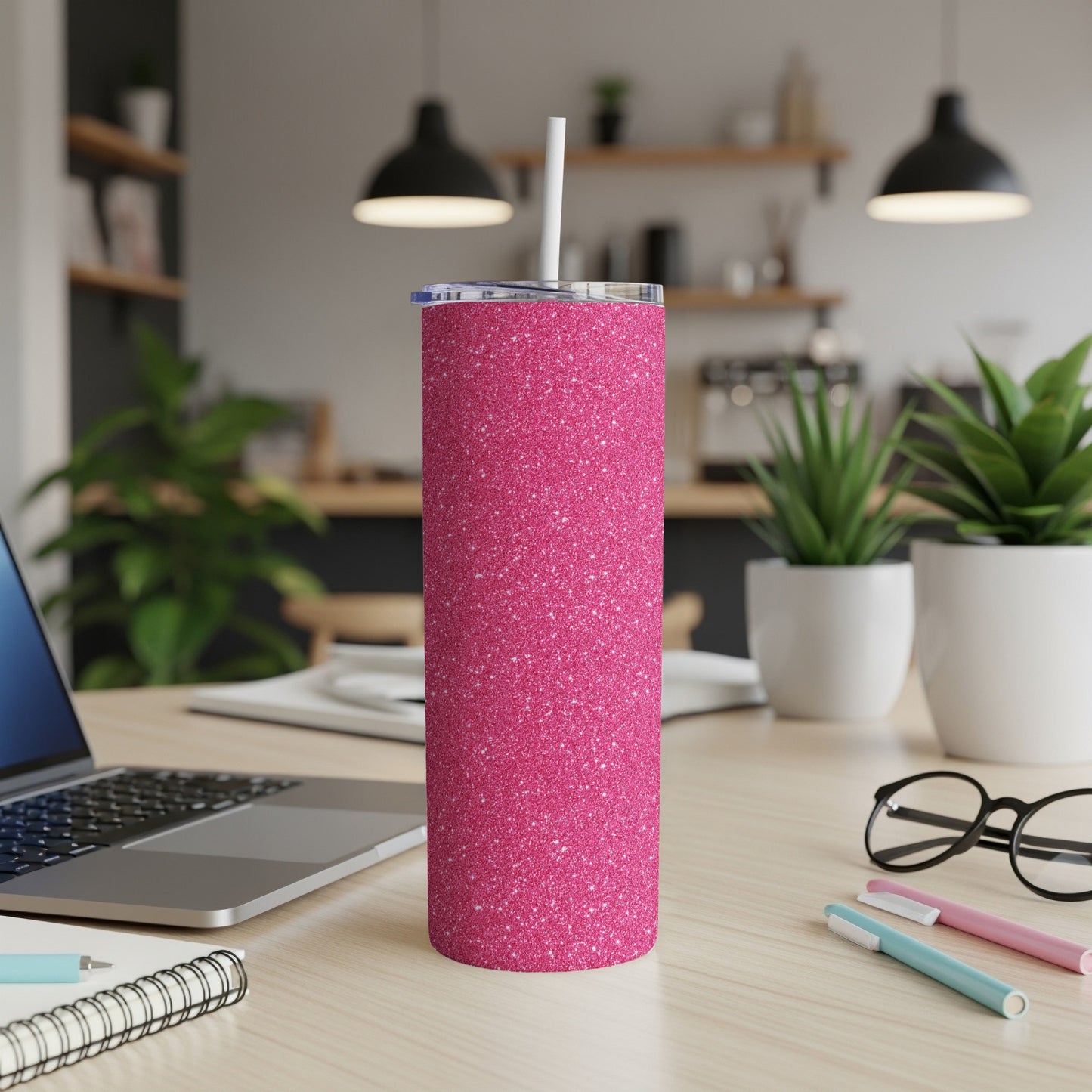 Glittery pink tumbler with a straw on a wooden desk next to a laptop and glasses.