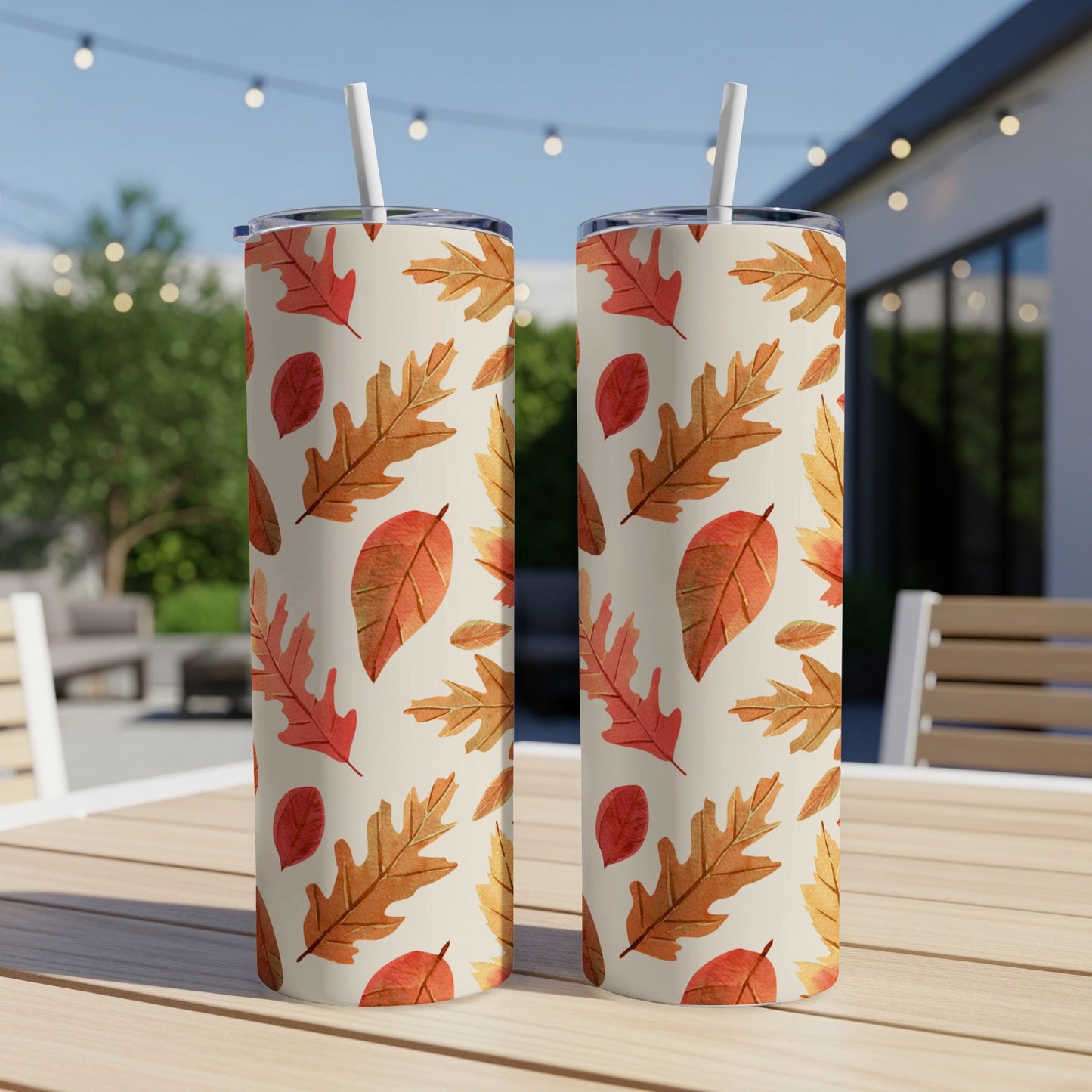 Two tall tumblers with a repeating pattern of autumn leaves on an outdoor table.