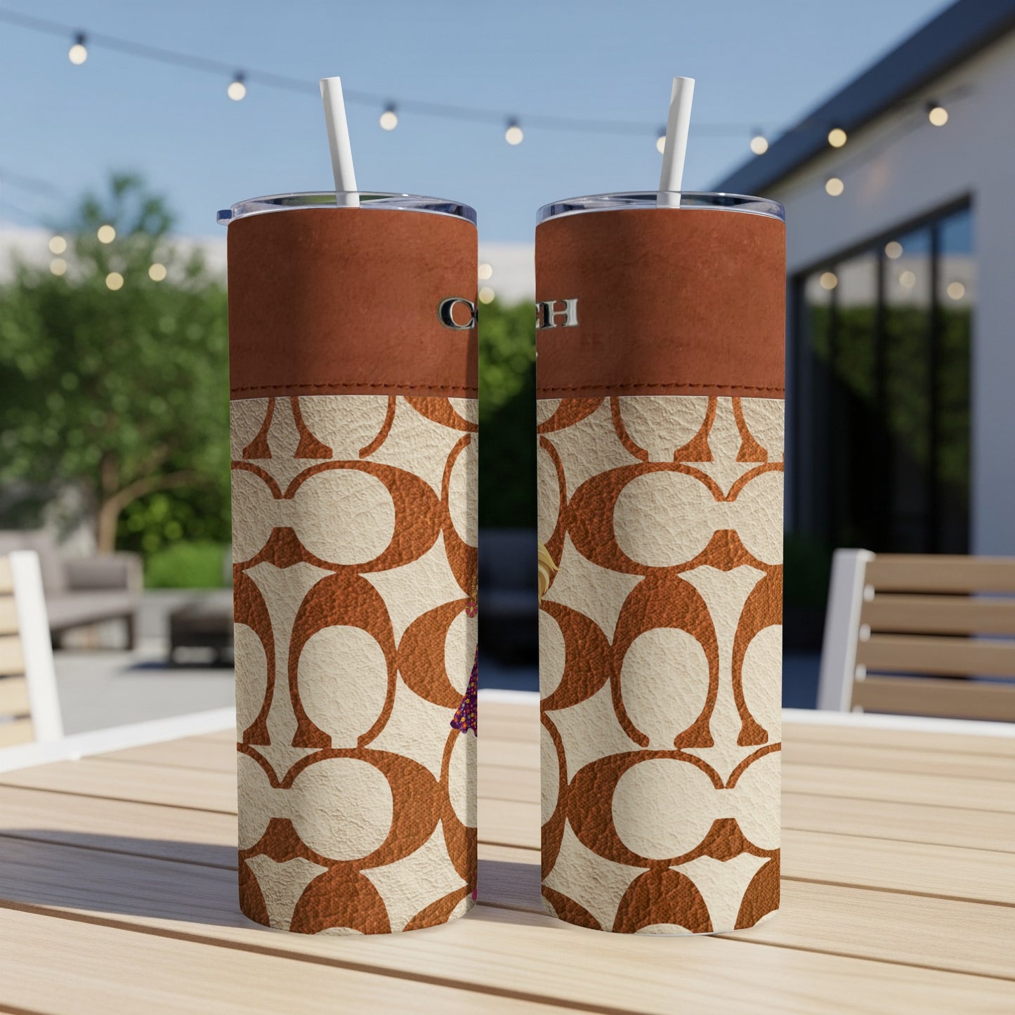 Two Coach print tumblers with straws on a wooden table outdoors.
